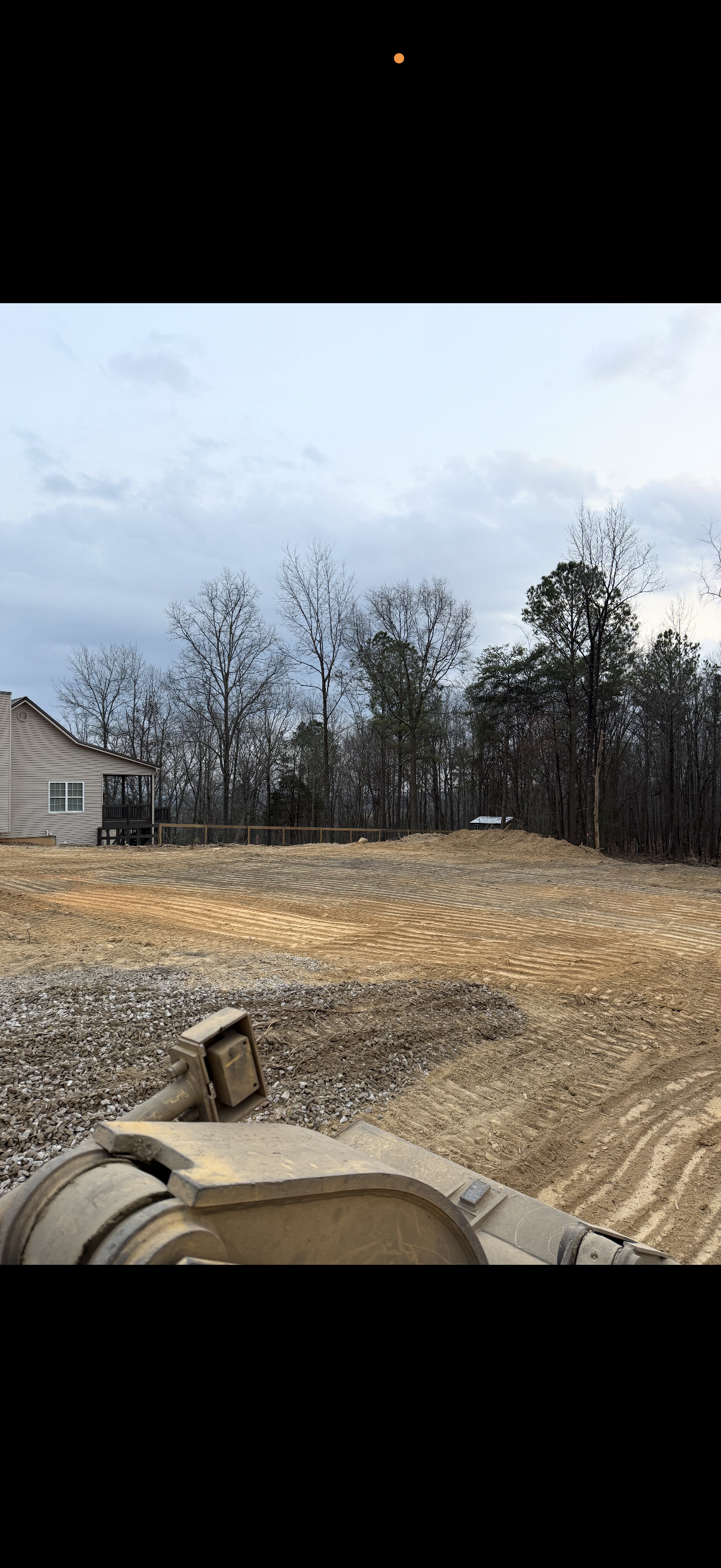 Construction site with cleared dirt and an excavator in foreground, a house on the left, and a wooded area with leafless trees in the background under a cloudy sky.