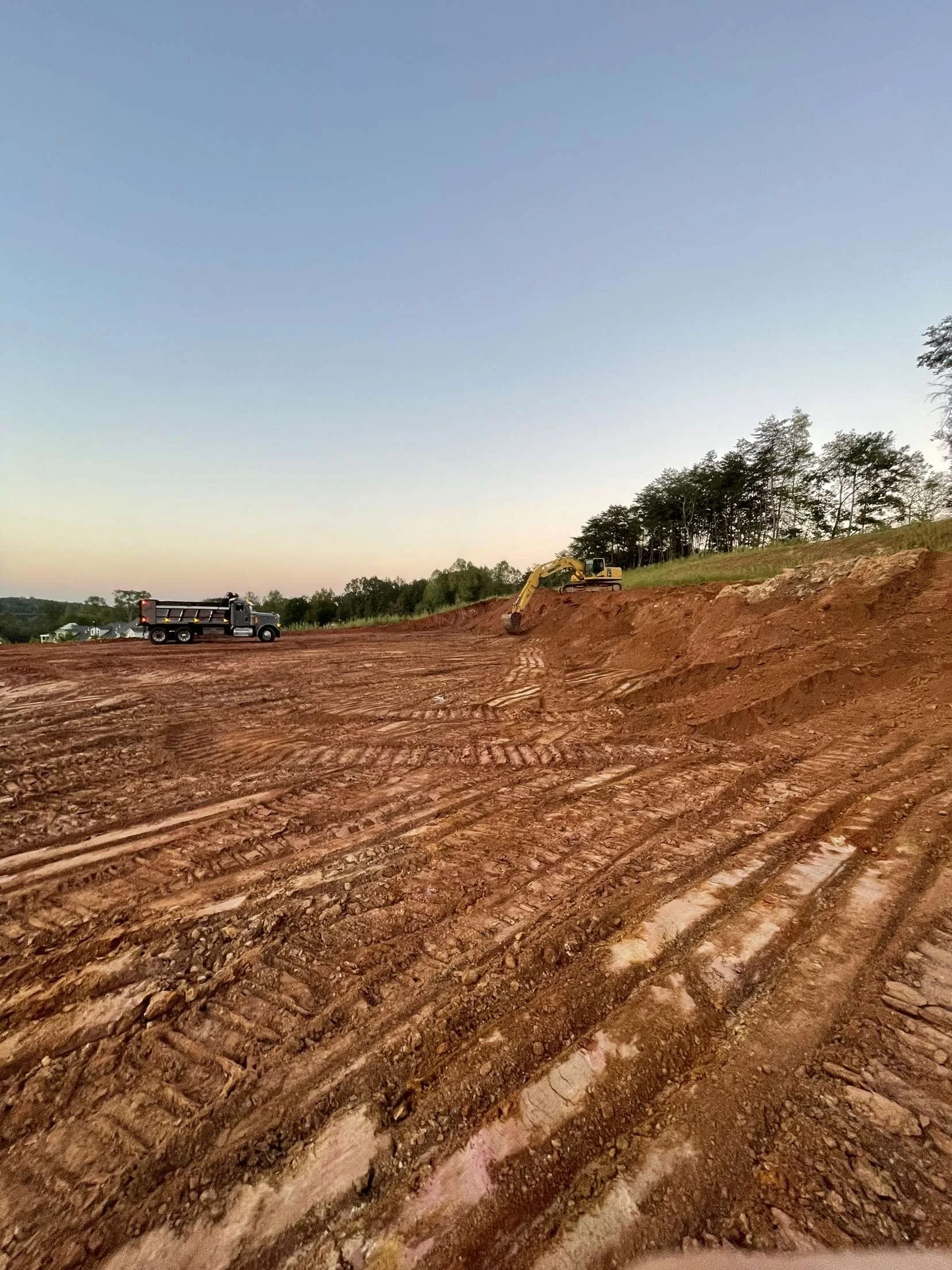 Construction site with dirt and tire tracks, a dump truck, a small excavator digging into the hillside, and trees in the background under a, clear sky at sunset.