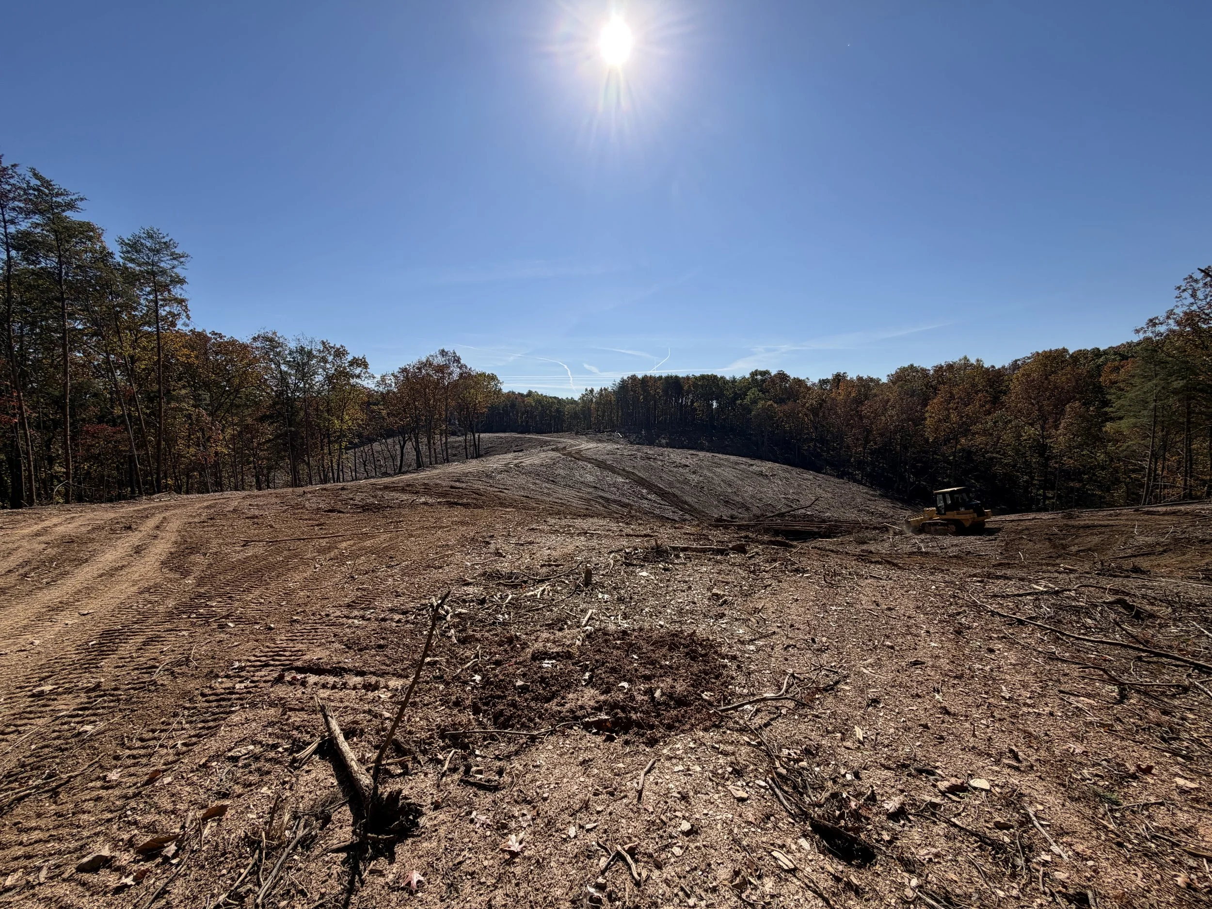 A cleared construction site with dirt and debris in the foreground, a small bulldozer on the right, and a forest of trees with changing leaves in the background under a clear blue sky with the sun shining.