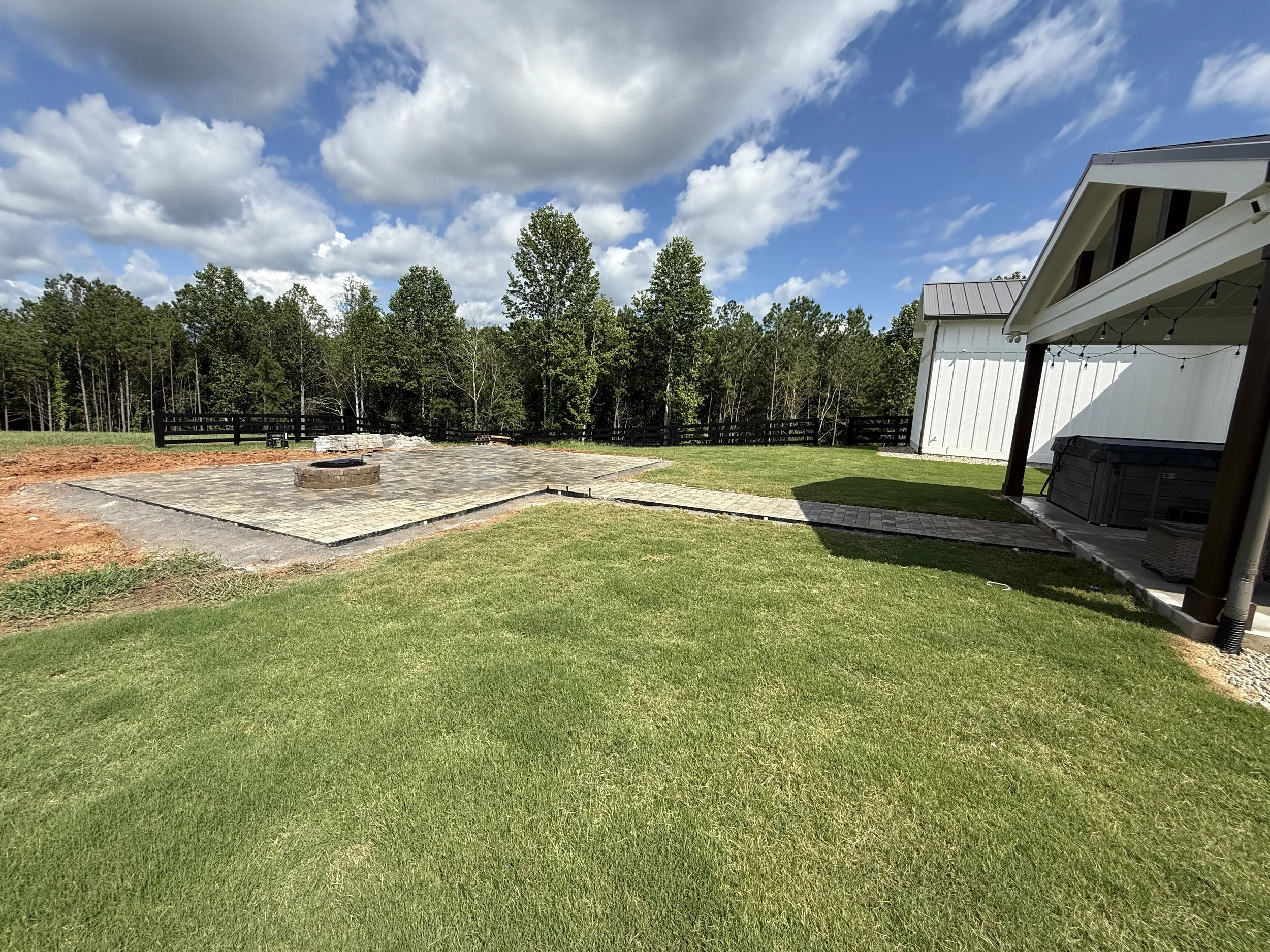 Backyard with green grass, a stone-paved patio area with a fire pit, a white shed, a hot tub, and a fence with trees in the background, under a partly cloudy sky.