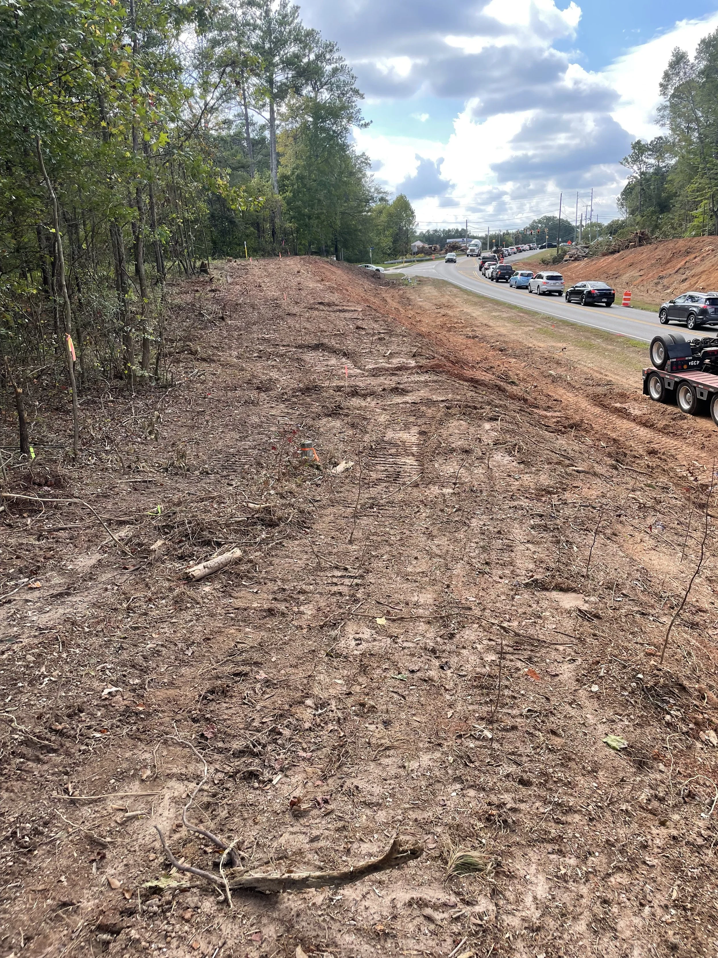Construction site along a highway with cleared dirt and a line of cars on the road. Green trees are on the left and right sides, and the sky is partly cloudy.
