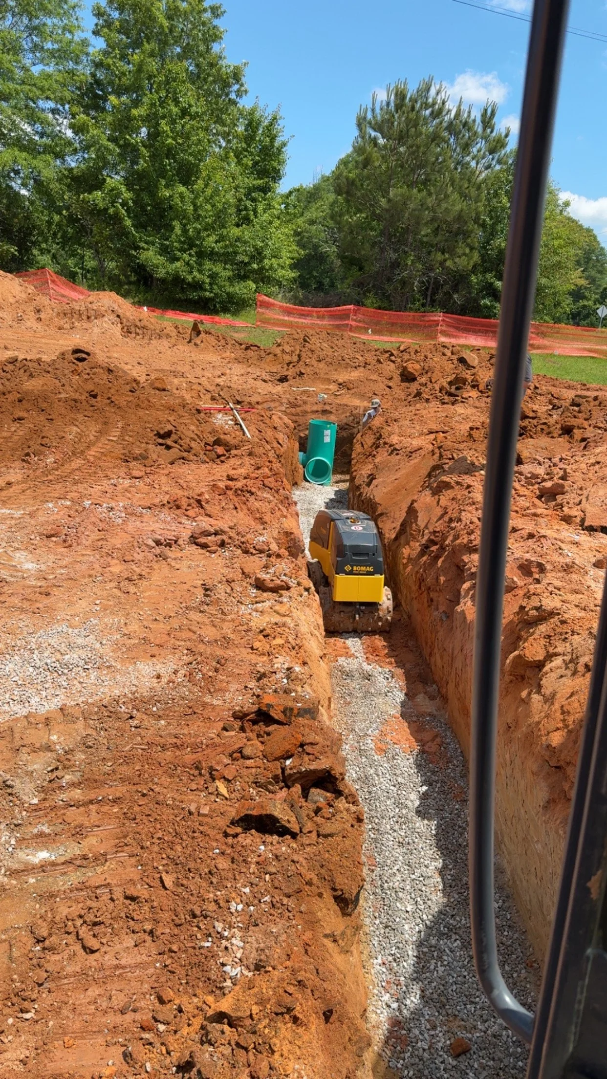 Construction site with trench being dug, a yellow compact excavator working in the trench, large dirt mounds on either side, green trees in the background, and a blue sky with some clouds.