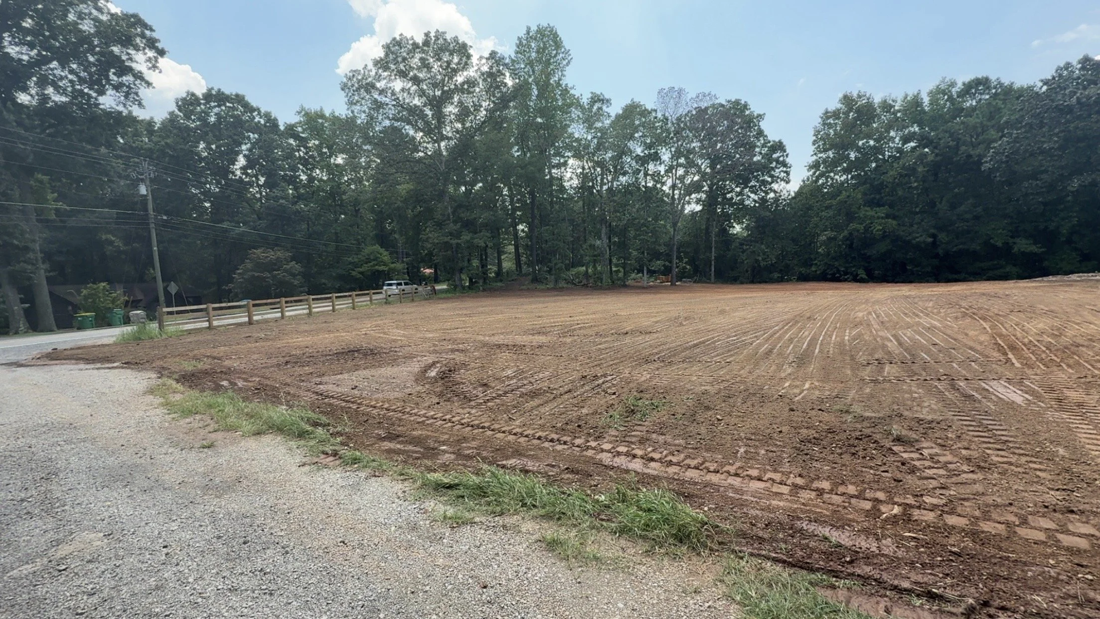 A cleared patch of land with tire tracks, next to a gravel road and a wooded area with tall trees in the background.