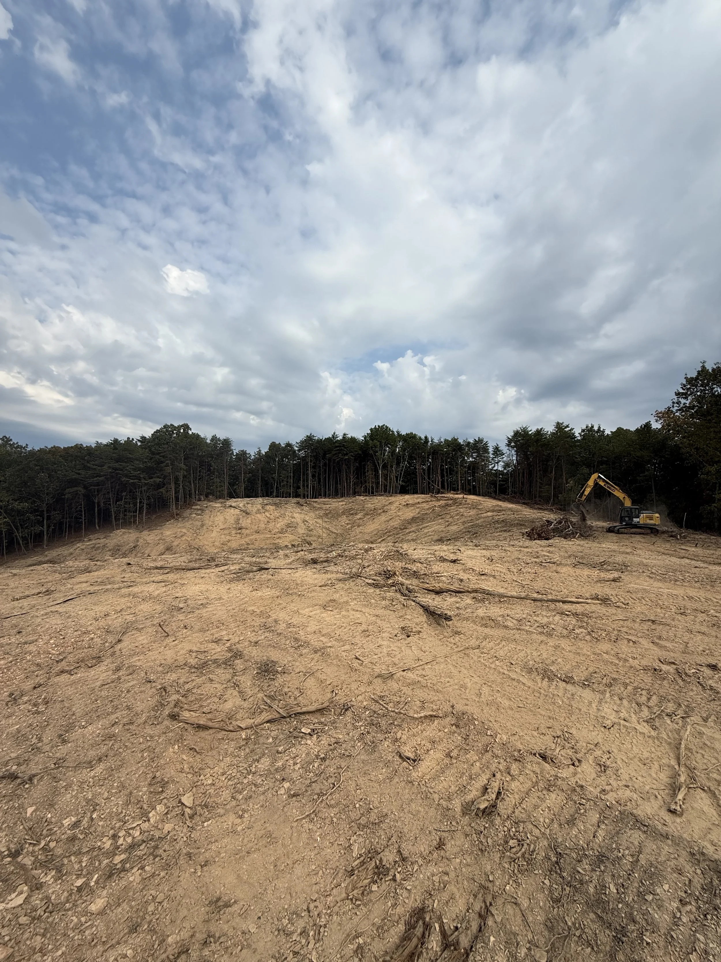 A construction site with a large area of bare dirt and an excavator working near a tree line under a cloudy sky.
