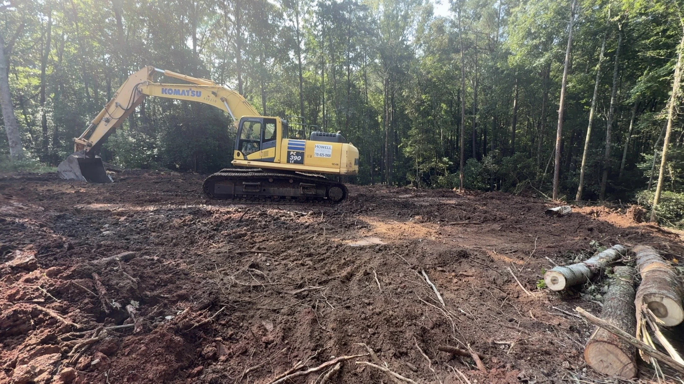 A yellow Komatsu excavator on a dirt clearing surrounded by trees, with logs on the ground nearby.