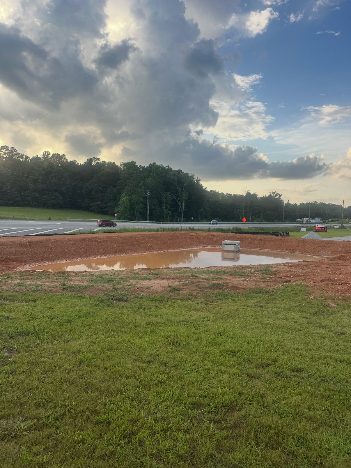 A small pond of water on a dirt patch near a grassy field, with a railroad crossing sign and a highway with cars passing by in the background under a partly cloudy sky.