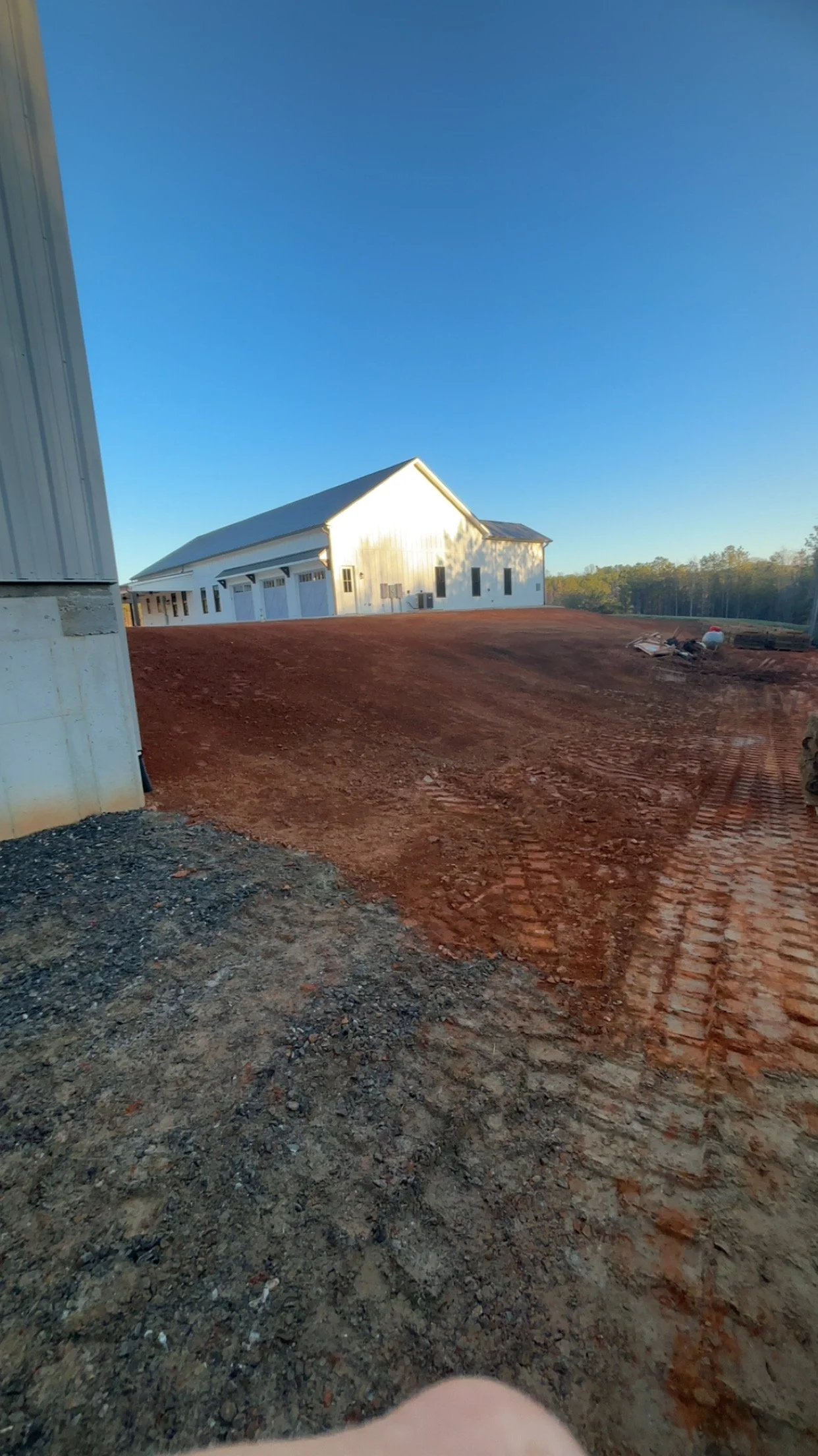 A rural construction site with a large, white barn-like structure and a dirt driveway under a clear blue sky.