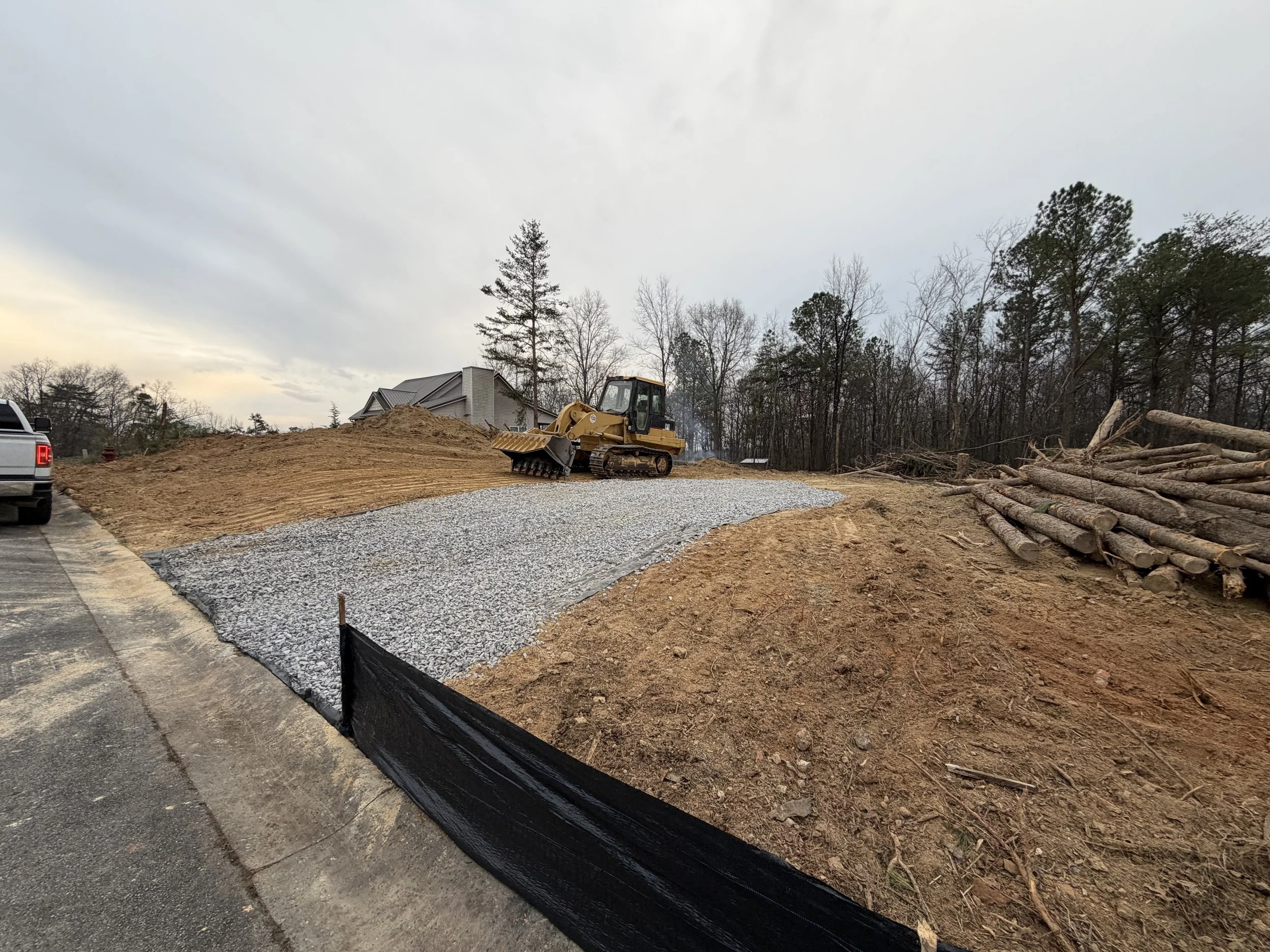 Construction site with a bulldozer on a gravel path, a pile of dirt, and stacked logs, with a house and trees in the background under a cloudy sky.
