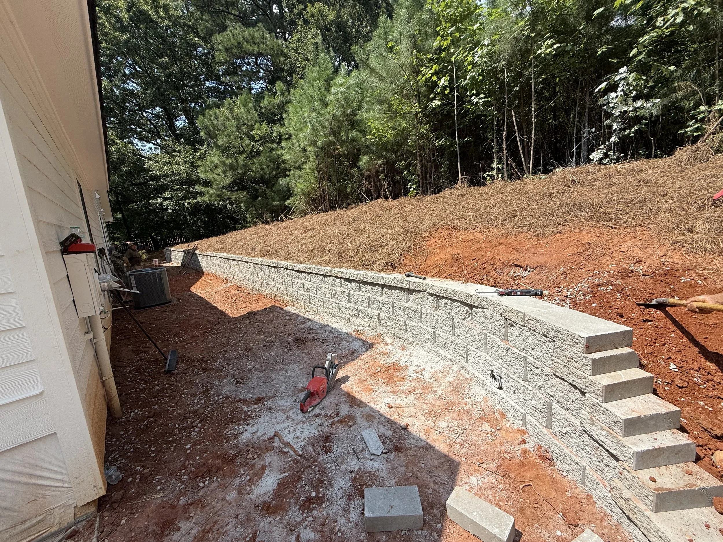 Construction site with a newly built concrete retaining wall beside a house, with tools and equipment lying on the ground and a wooded area in the background.