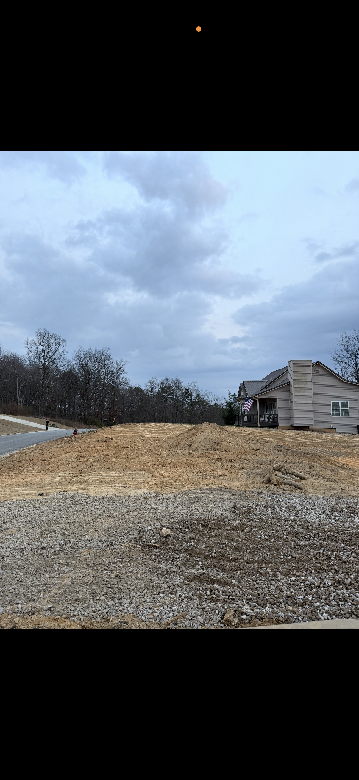 A residential area under construction with a house on the right, dirt piles, and a gravel-covered ground in the foreground.
