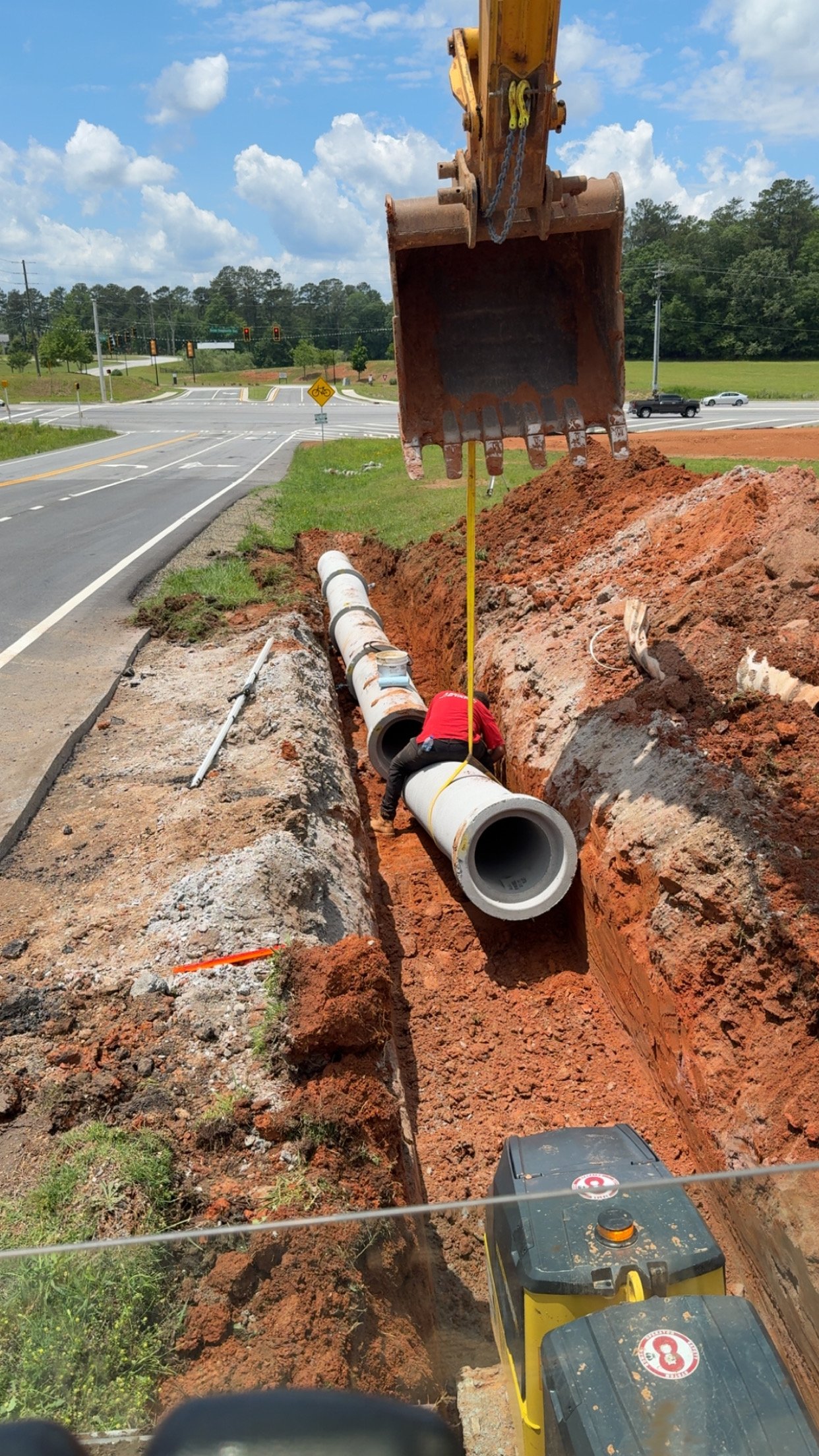 Construction worker installing large pipes in a trench along a roadside, with orange soil and construction equipment visible.