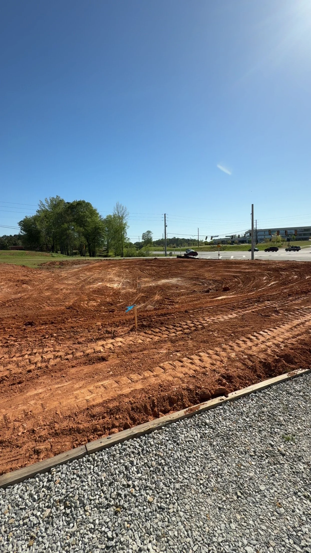 Construction site with truck tracks on red dirt, a small blue flag marker, and a gravel path in the foreground, with a busy road, trees, and building in the background under a clear blue sky.