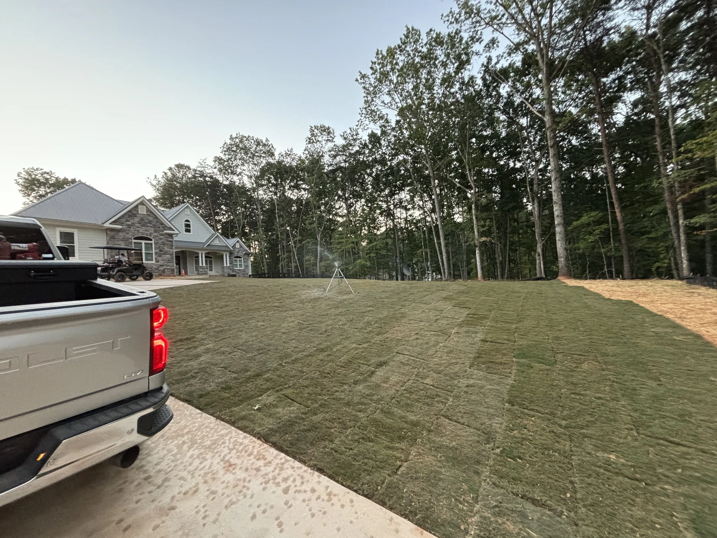 Lawn being prepared for sod installation in front of a modern house, with installed unrolled sod patches visible, and truck parked nearby.