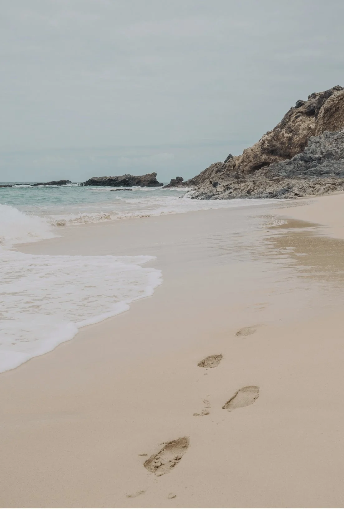 Footprints in the sand on a beach near rocks and gentle waves under a cloudy sky.
