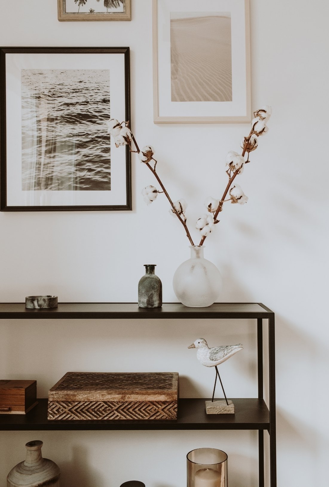 A wall decorated with framed beach photographs, a cotton branch in a white vase, and decorative objects including small vases and a bird sculpture on a black shelf.