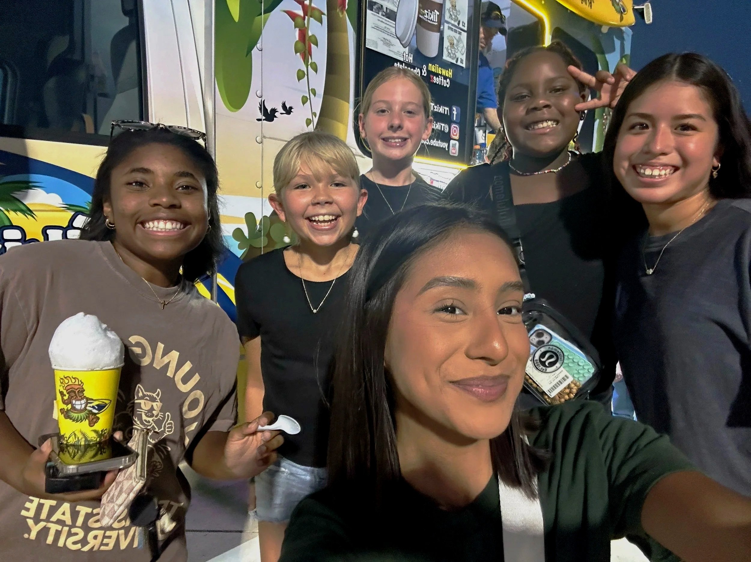 Group of smiling children taking a selfie in front of a food truck at night, with one girl holding a tasty-looking vanilla soft serve ice cream cone.