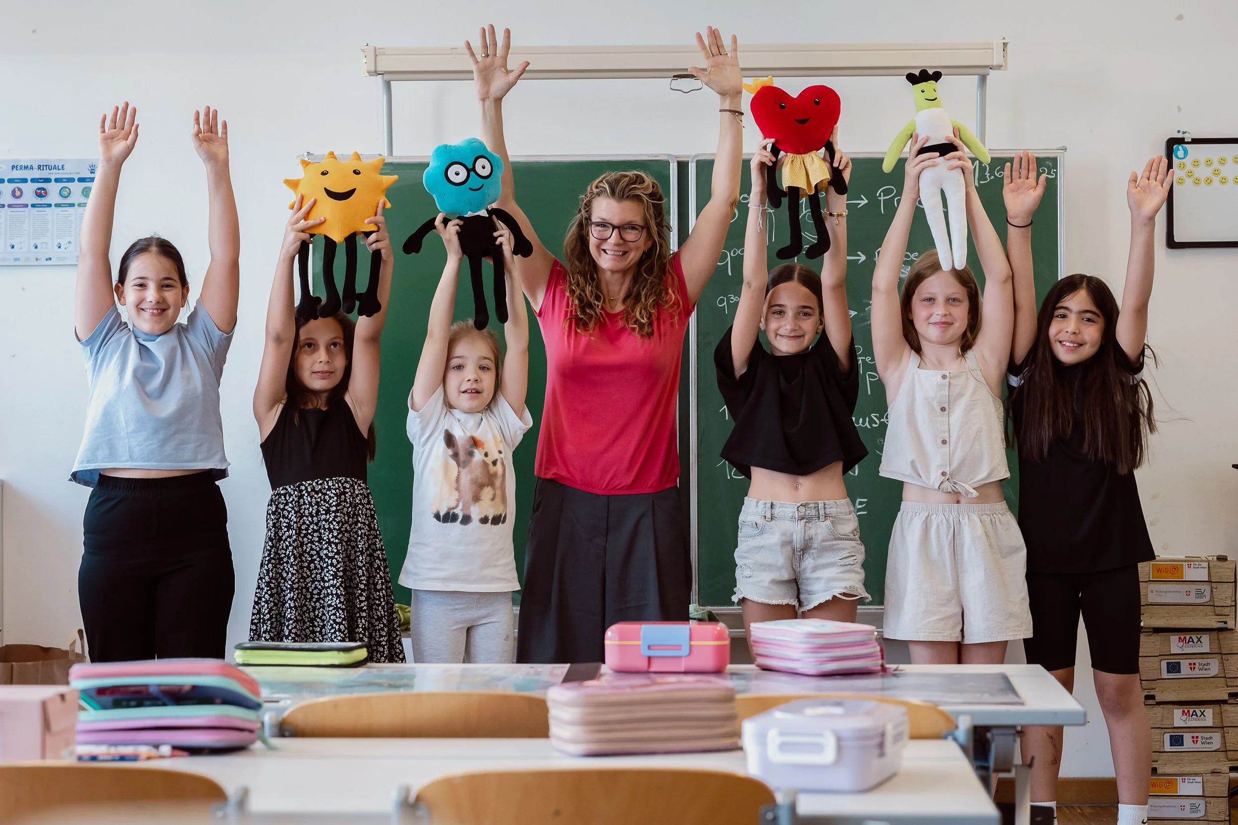 Eine Lehrerin mit sechs Schülern in einem Klassenzimmer, die kreative Handpuppen hält. Alle lächeln und stehen vor einer Tafel.