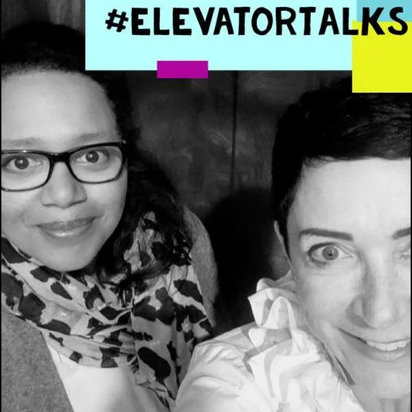 Zwei Frauen lächeln, eine trägt eine Brille und ein Muster-Schal, die andere hat kurze dunkle Haare und trägt eine weiße Bluse. Im Hintergrund ein heller, stilisierter Text mit #ELEVATORTALKS auf einem hellblauen Hintergrund, ergänzt mit farbigen Akzenten.