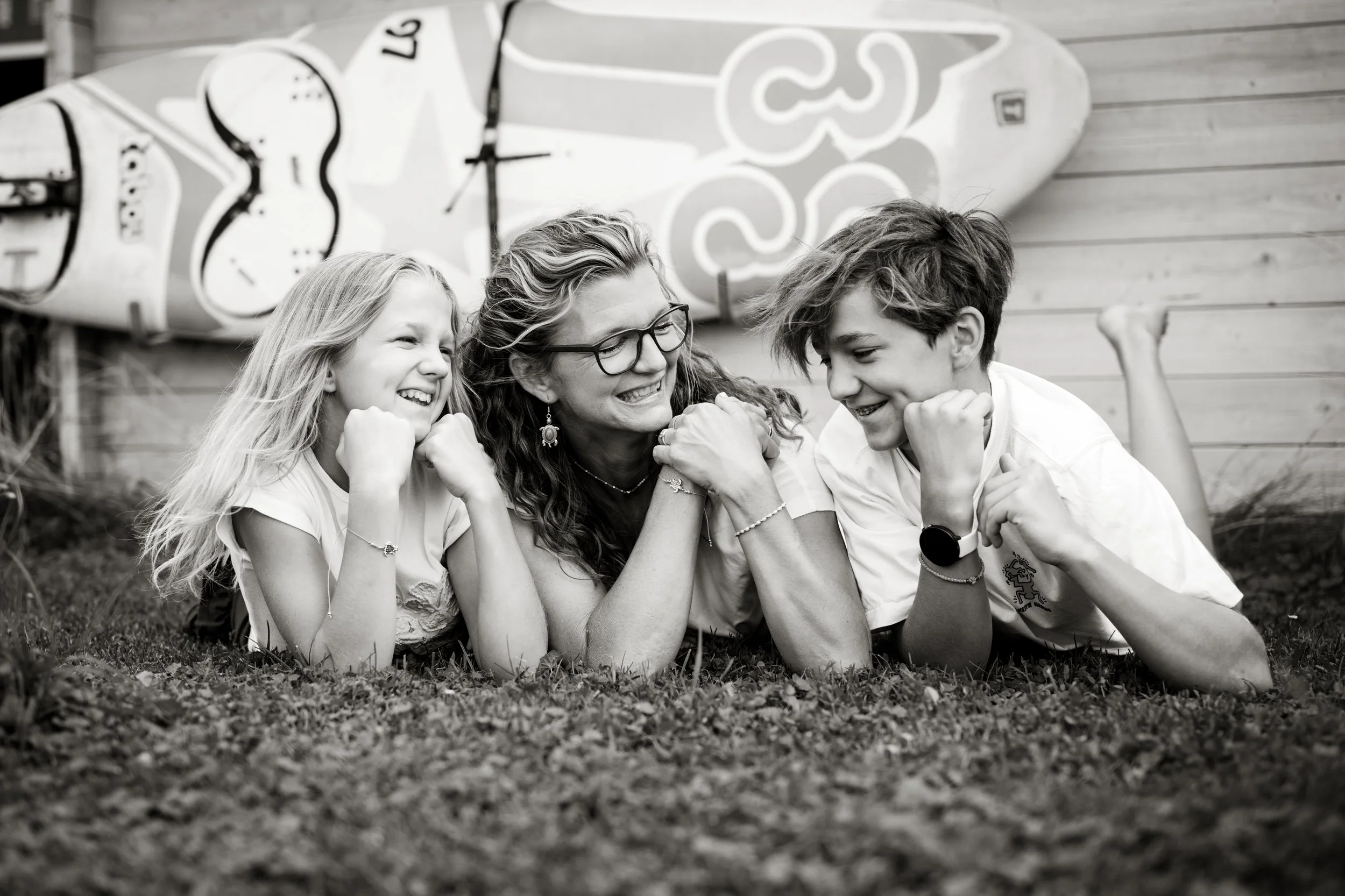 Ein Mutter und ihre zwei Kinder lachen lachend auf dem Boden im Garten, mit einem Surfbrett im Hintergrund.