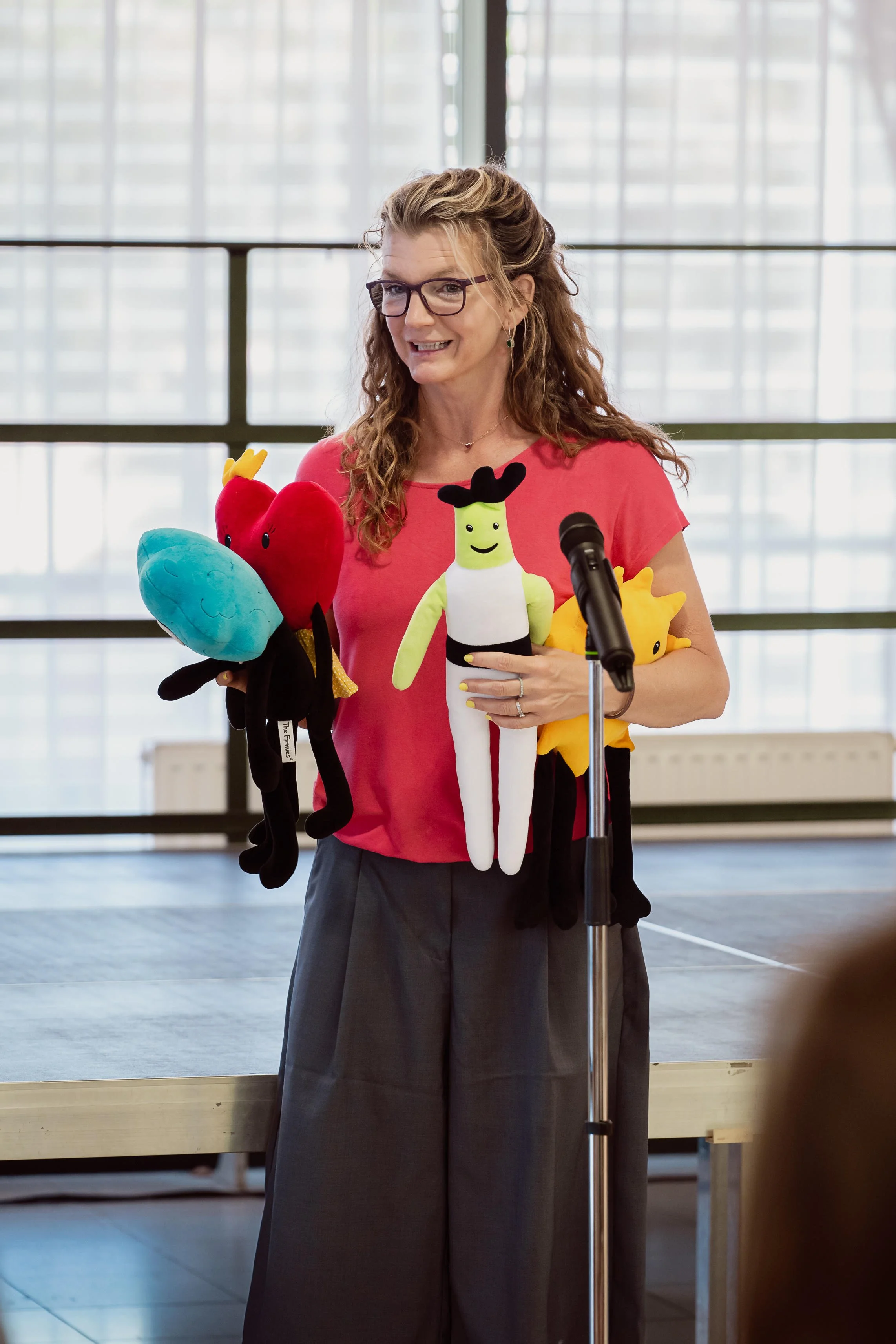 Frau mit Brille und lockigem Haar hält Puppets in einer Halle, im Hintergrund große Fenster.