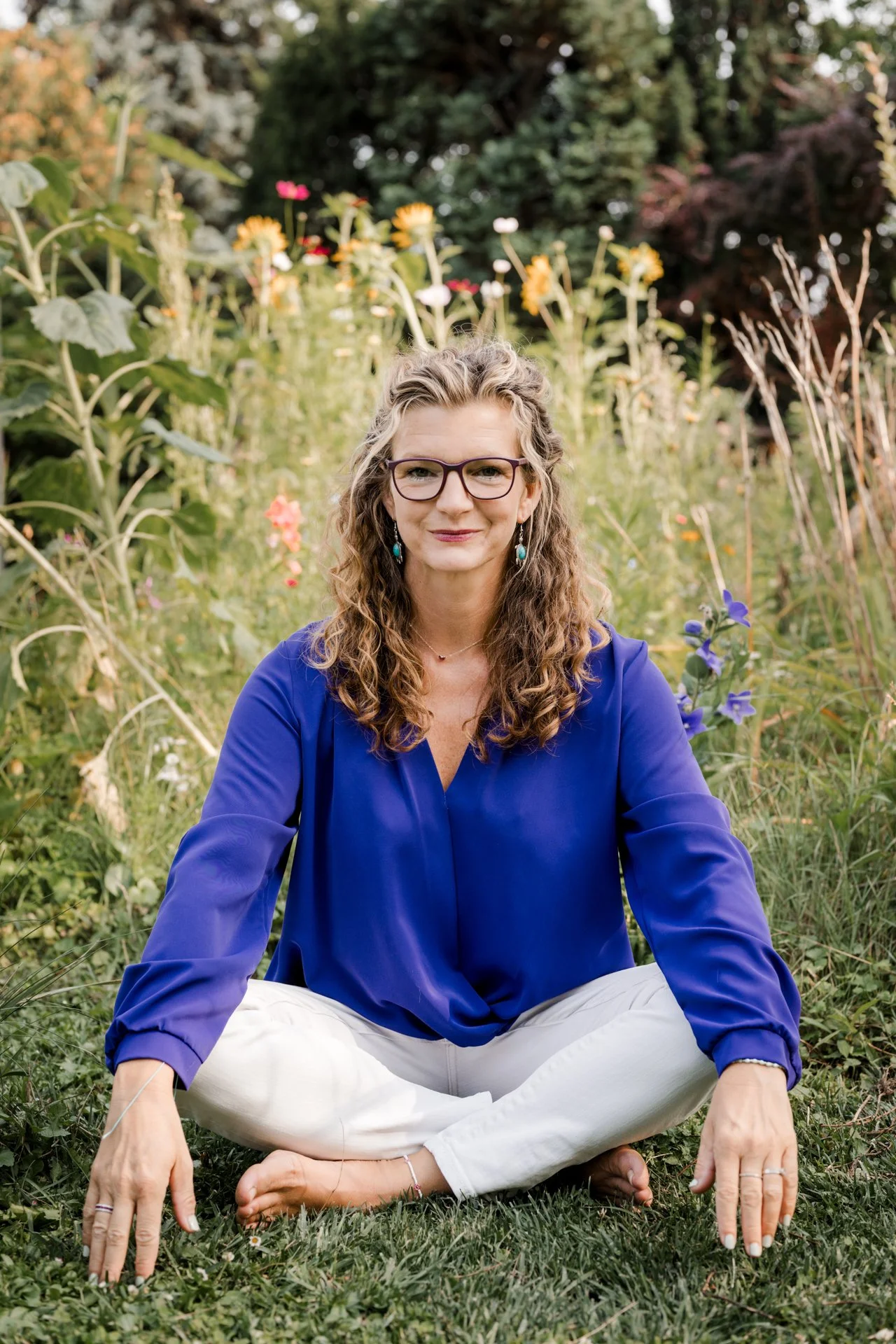 Eine Frau sitzt in einem Garten mit bunten Blumen, trägt eine blaue Bluse, weiße Hosen, Brille und Ohrringe, und lächelt freundlich.