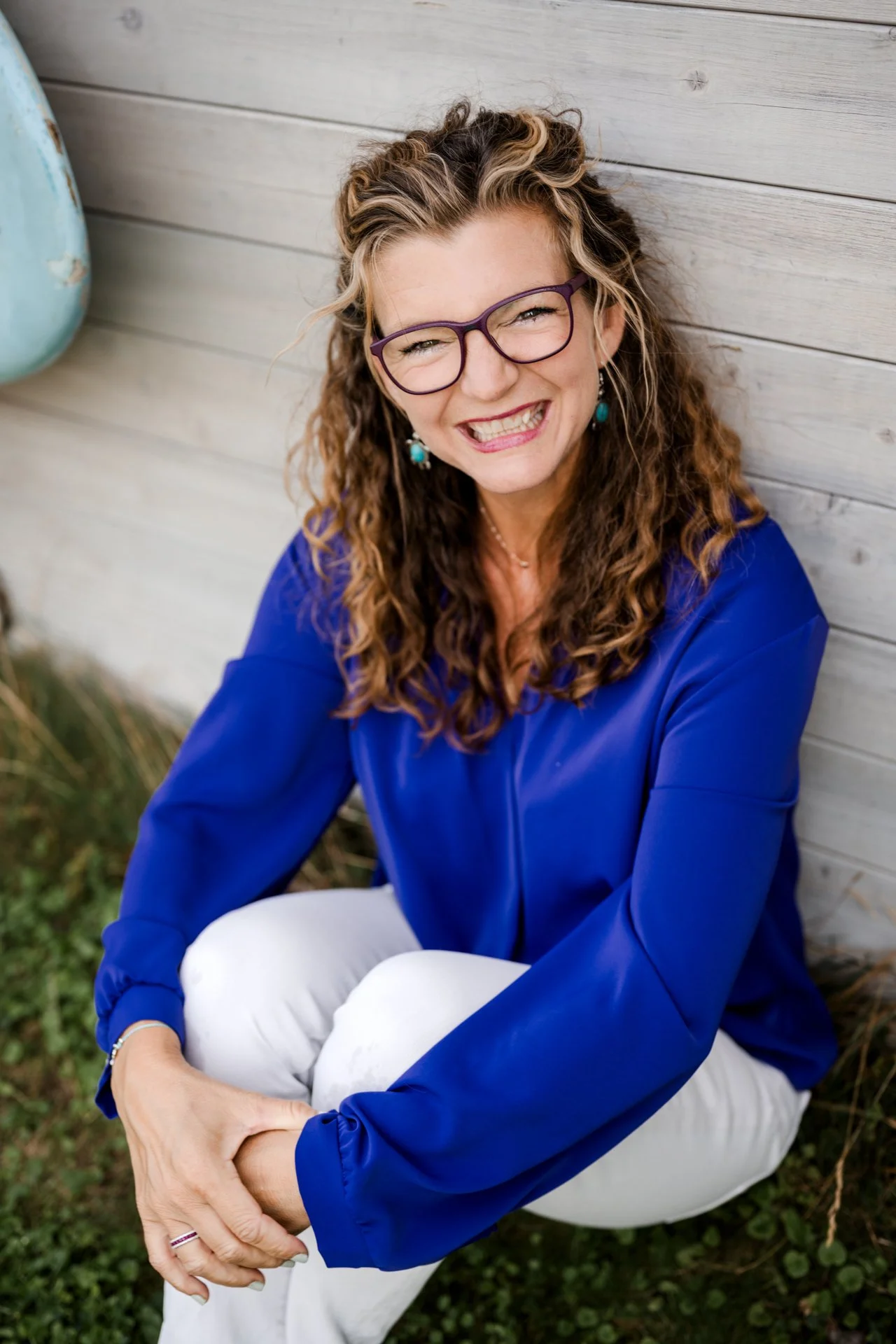 Eine lachende Frau mit Brille, braunen lockigen Haaren, blauer Bluse und weißen Hosen sitzt vor einer Holzwand.