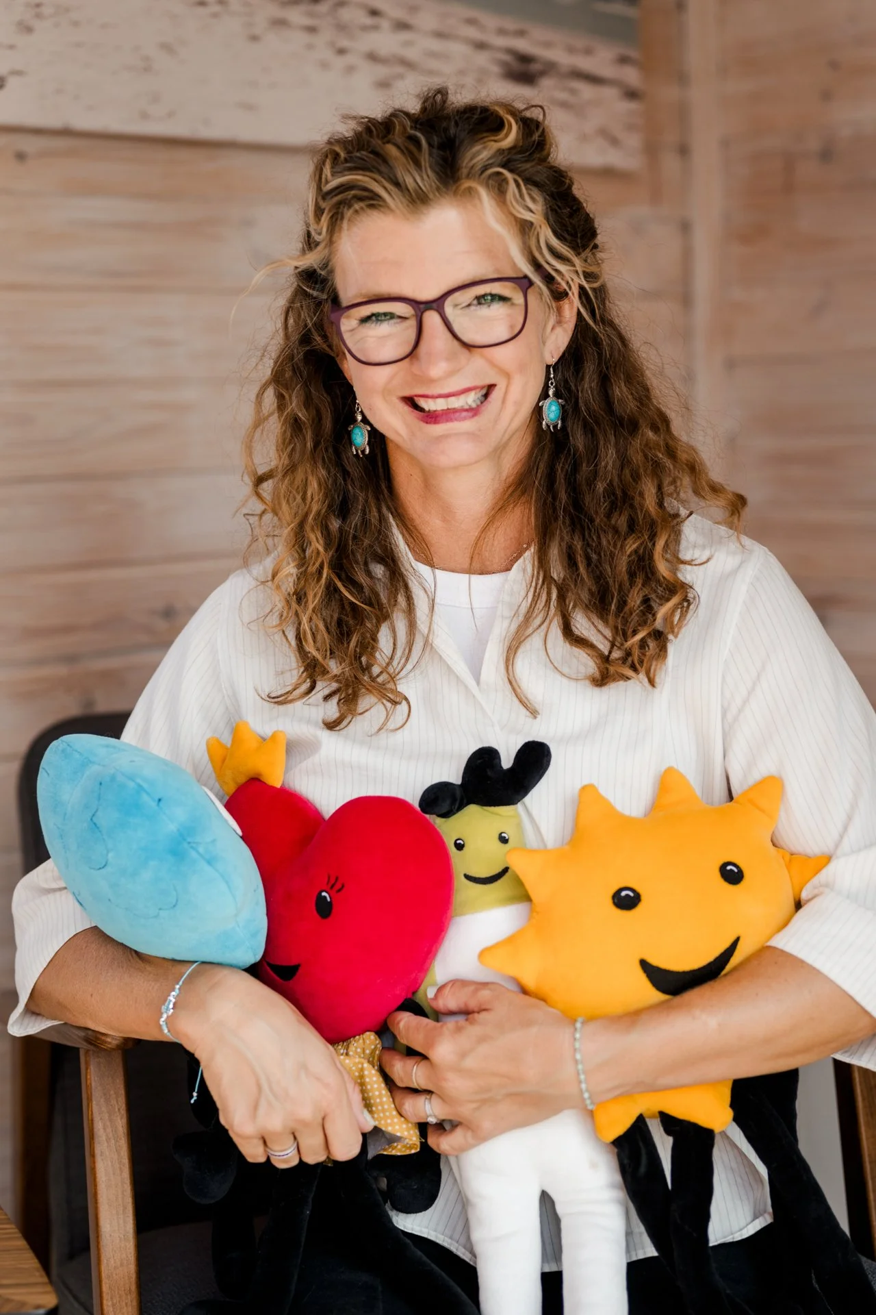 Eine lachende Frau mit brünetten, lockigen Haaren und Brille hält mehrere bunte Plüsch-Kuscheltiere, darunter einen blauen, einen roten, einen schwarzen, einen weißen und einen gelben Sonnenschein, vor einer holzvertäfelten Wand.