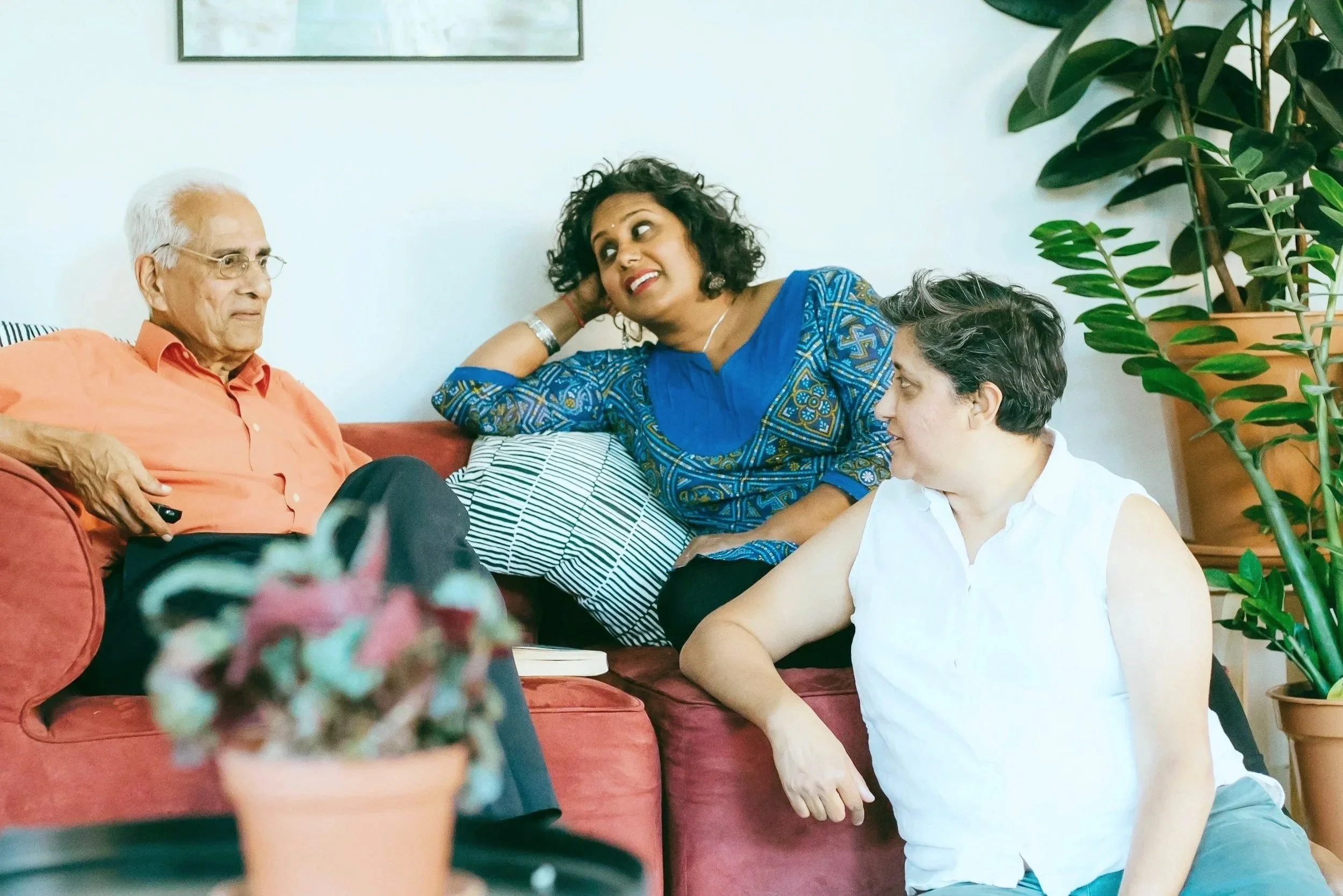 Four people sitting in a room having a conversation. An older man in an orange shirt on a red couch, a woman in a blue patterned top leaning on the couch, and a woman in a white sleeveless shirt leaning on the floor.