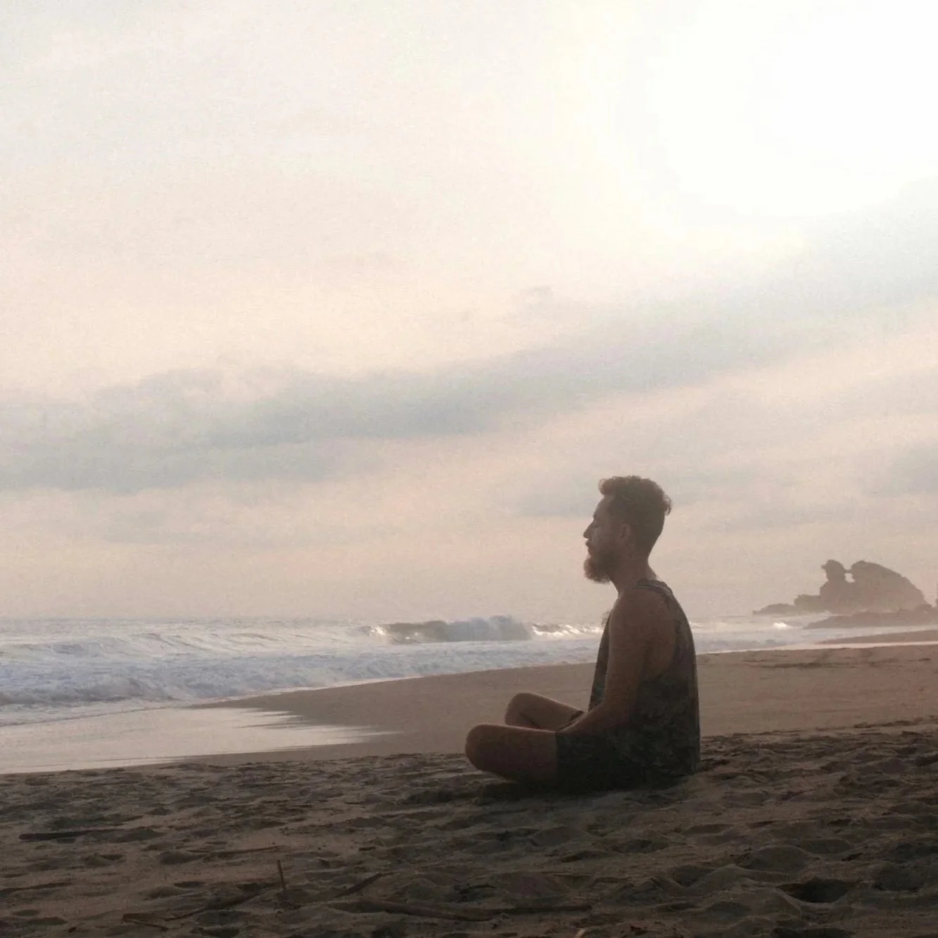 A man sitting cross-legged on the sand at the beach, facing the ocean during sunset or sunrise with a cloudy sky and rocky formations in the distance.