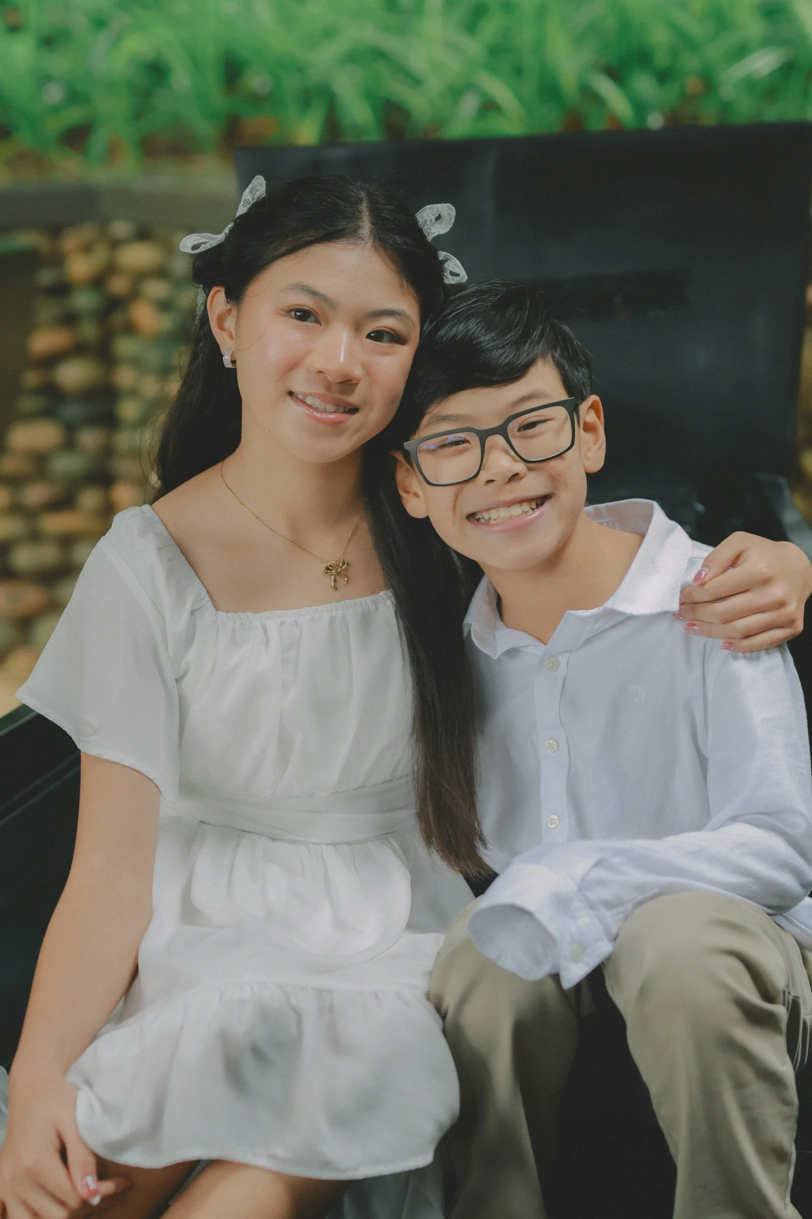 A woman and a boy smiling and sitting together, with the woman wearing a white dress and the boy wearing glasses and a white shirt, in front of a blurred, outdoor background.