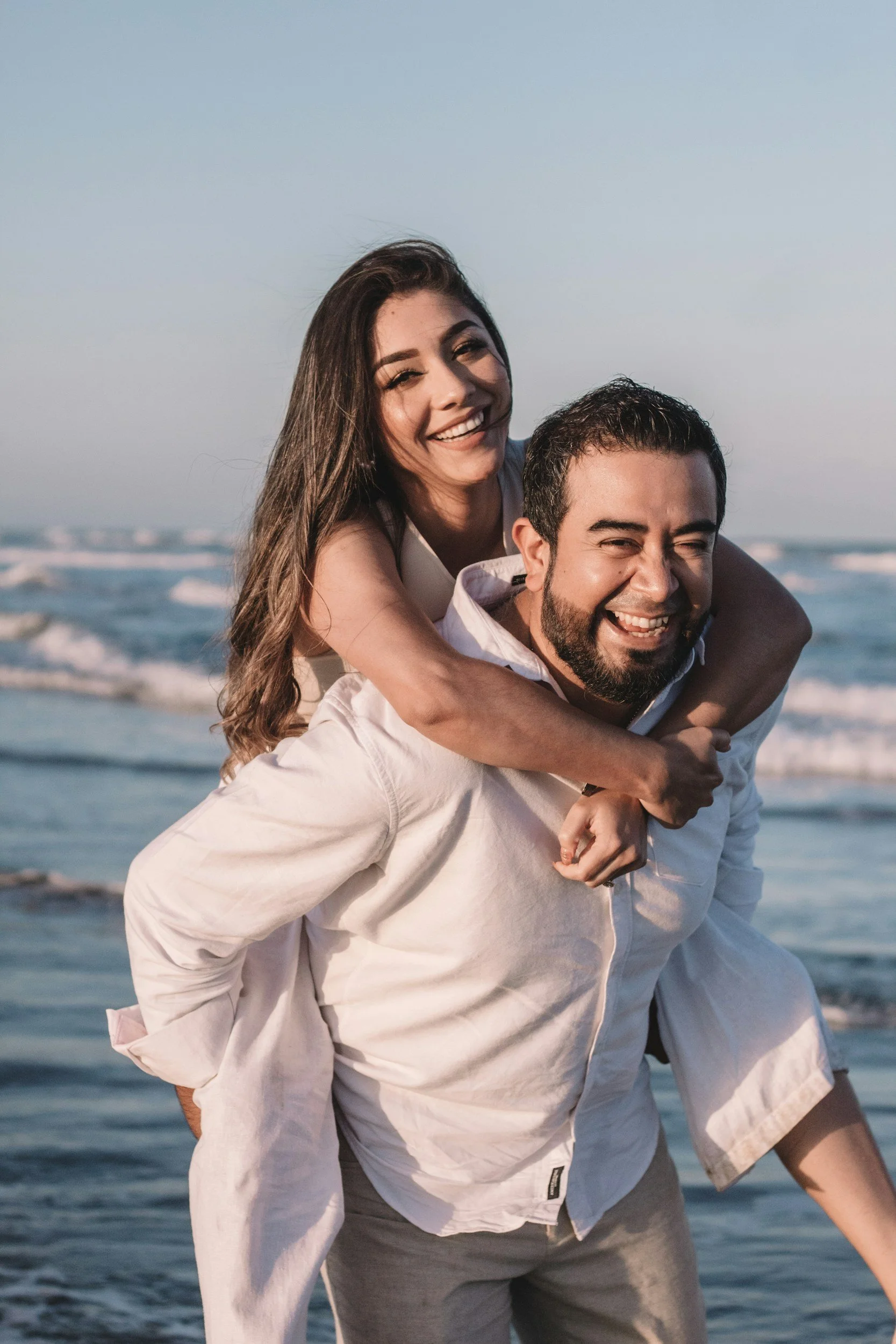 A happy couple at the beach, with the woman on the man's back, both smiling and enjoying the moment during sunset.