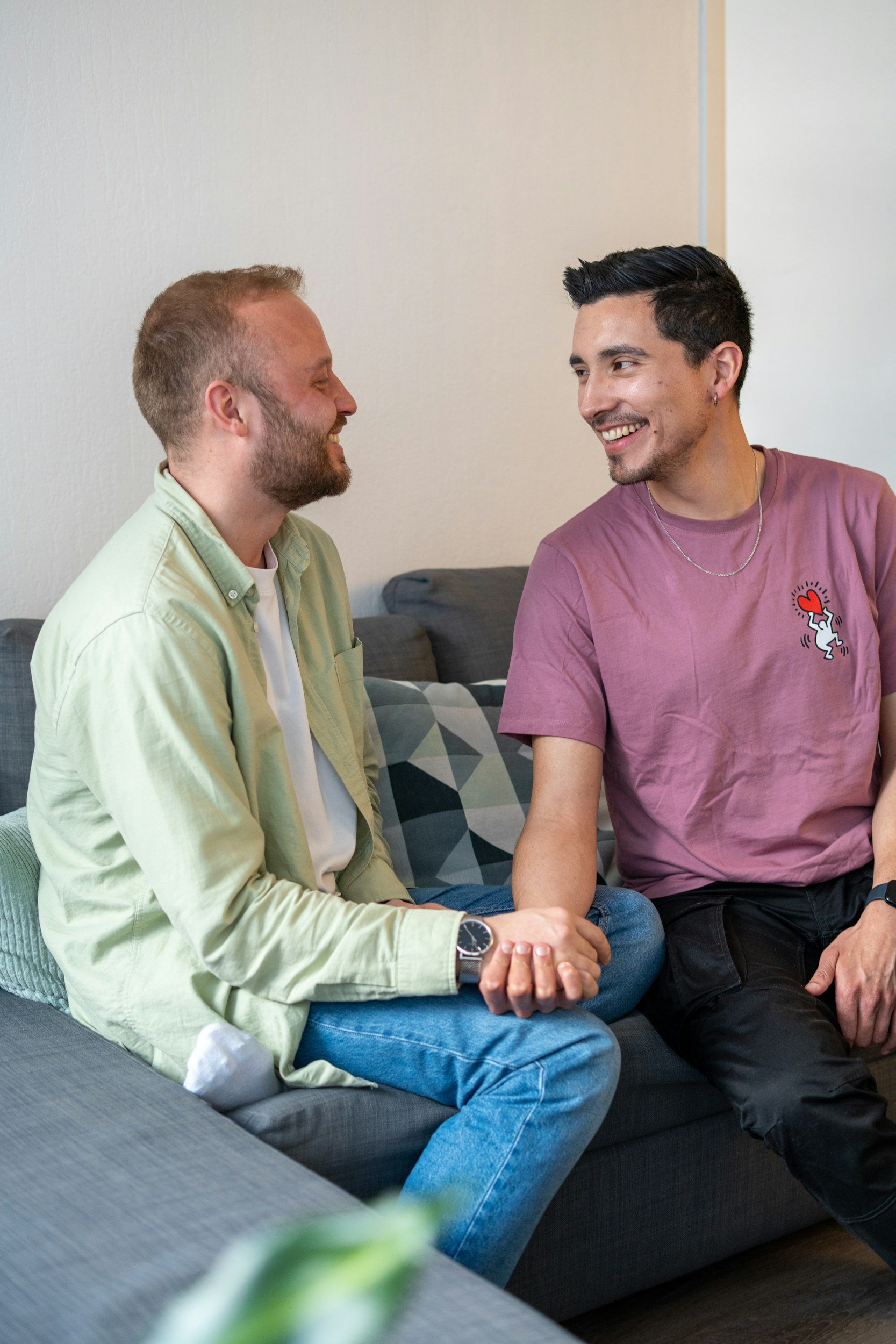 Two men sitting on a gray couch, smiling, holding hands, and looking at each other.