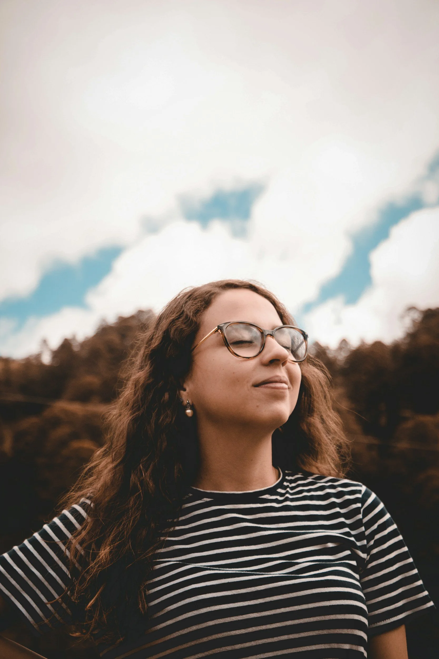 A woman with long wavy brown hair, wearing glasses and earrings, is smiling with her eyes closed. She is outdoors with a blurry background of trees and bushes, wearing a black and white striped shirt. The sky is partly cloudy.