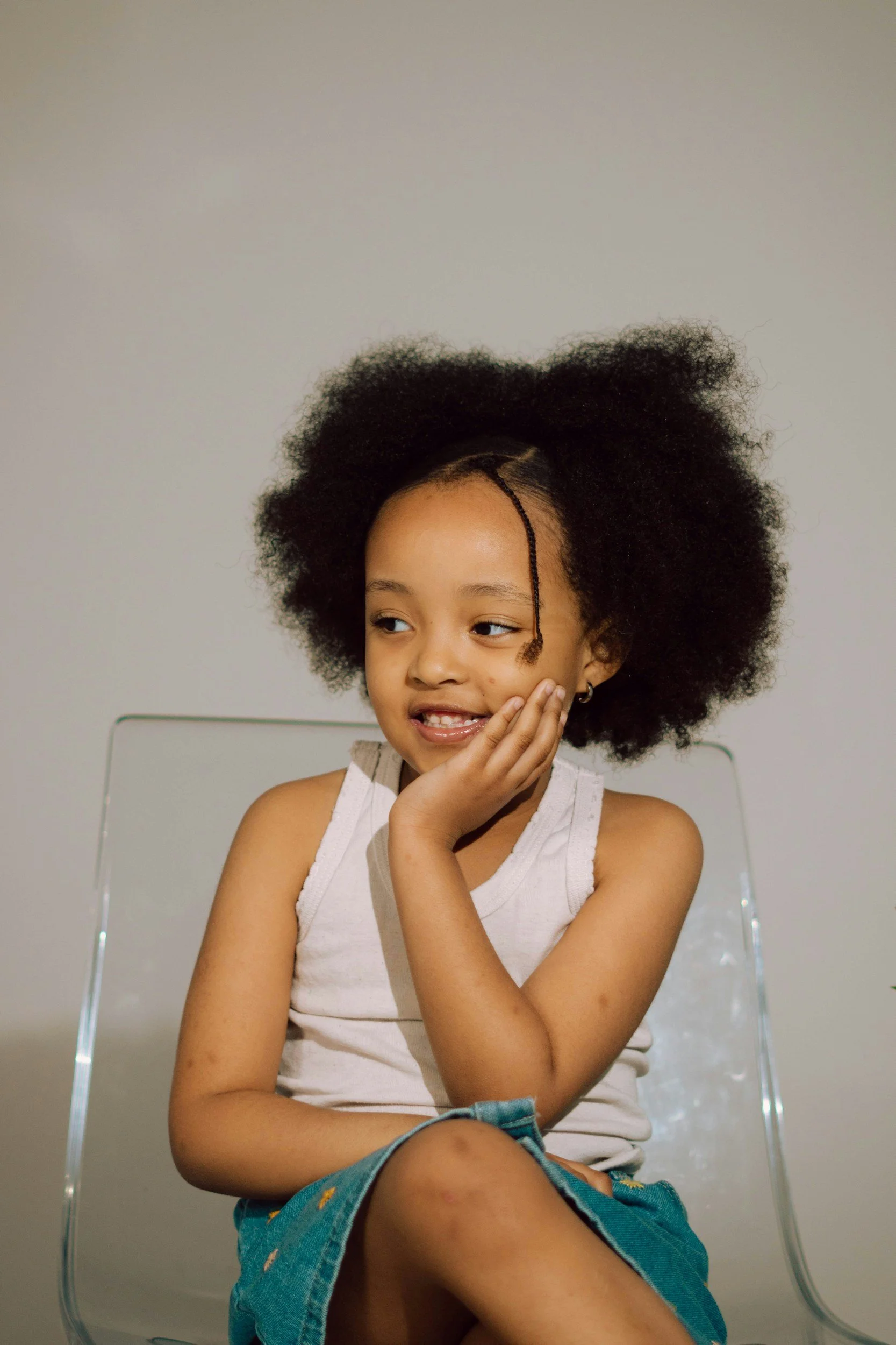 A young girl with natural curly hair sitting on a clear acrylic chair against a plain light-colored wall. She is resting her chin on her hand, smiling slightly, and looking to the side.