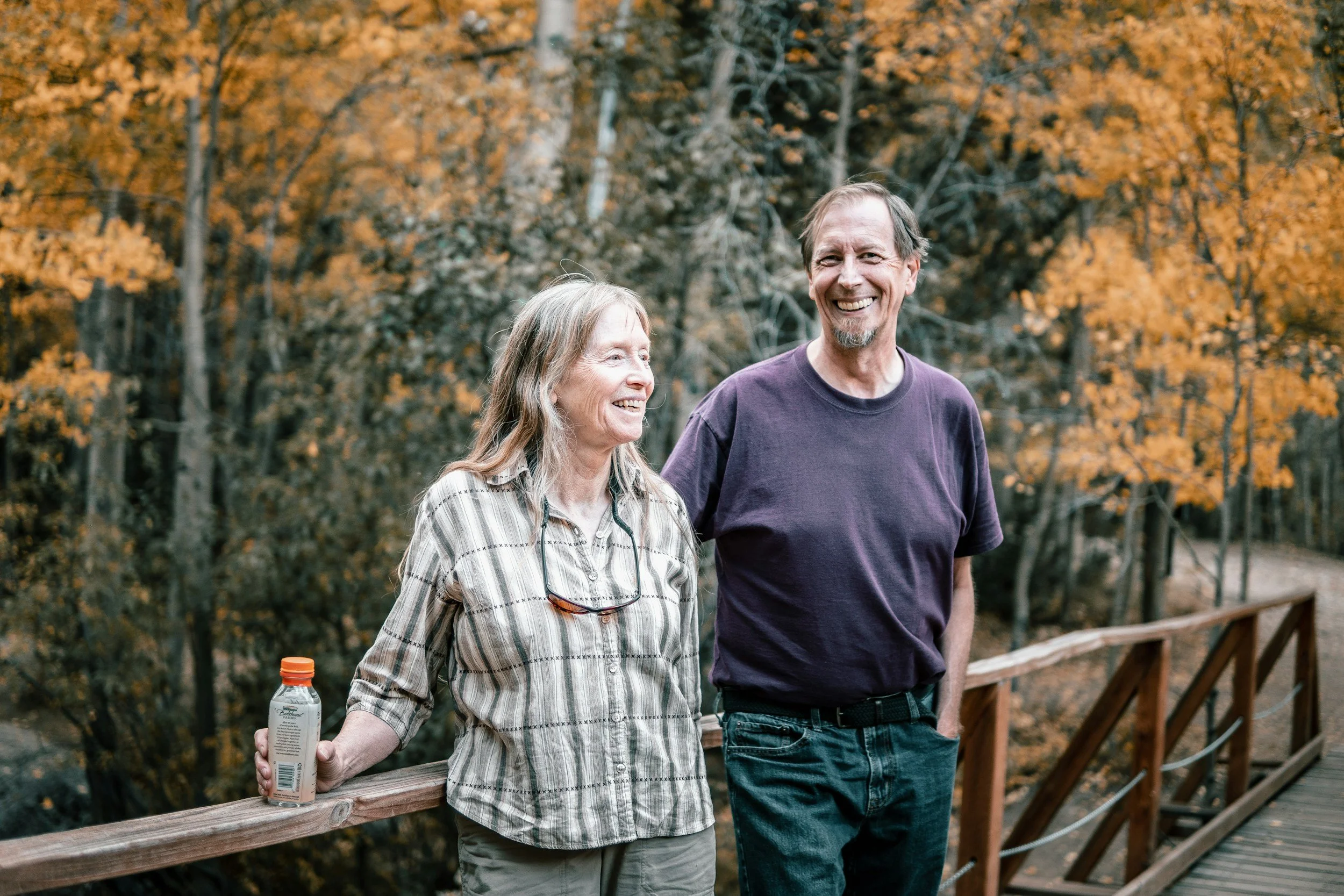 A smiling elderly woman and a smiling middle-aged man walk together on a wooden bridge in an autumn forest. The woman is holding a bottled drink and has glasses hanging around her neck. The man has a beard and is wearing a purple T-shirt.
