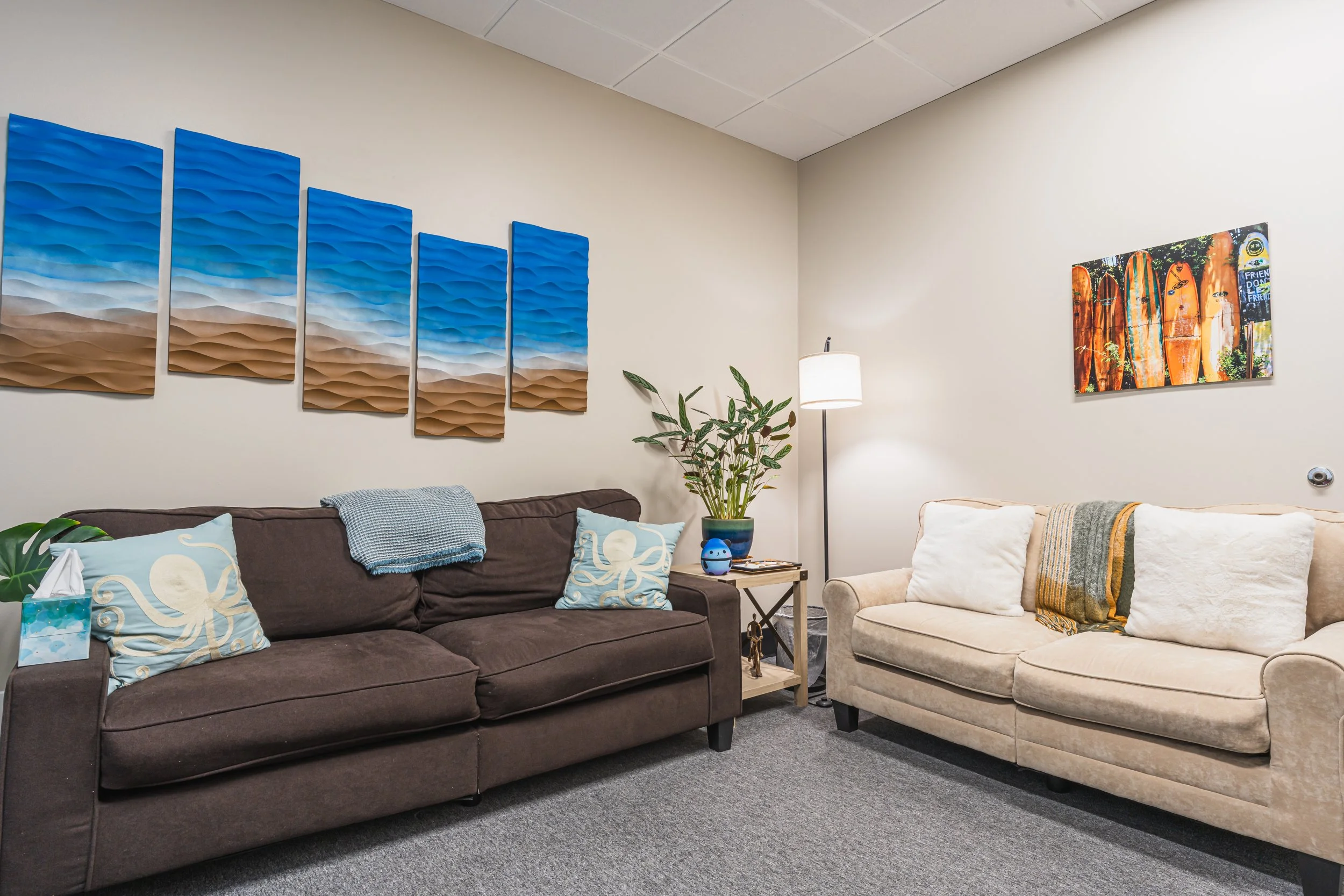 Living room with a brown couch, beige armchair, wall art, a plant, a lamp, and various pillows.