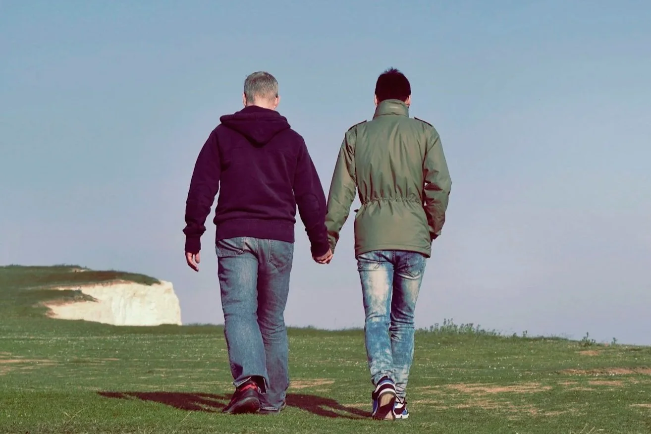 Two people walking hand in hand on a grassy field with a cloudy sky and white cliffs in the background