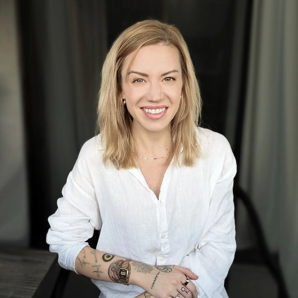 A woman with blonde hair smiling, wearing a white shirt, seated outdoors with a black background.