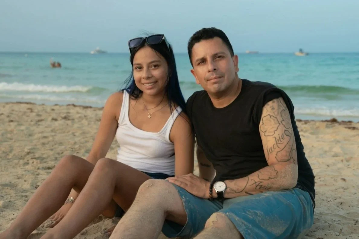 A man and woman sitting on a sandy beach near the ocean, with boats in the distance, smiling at the camera.