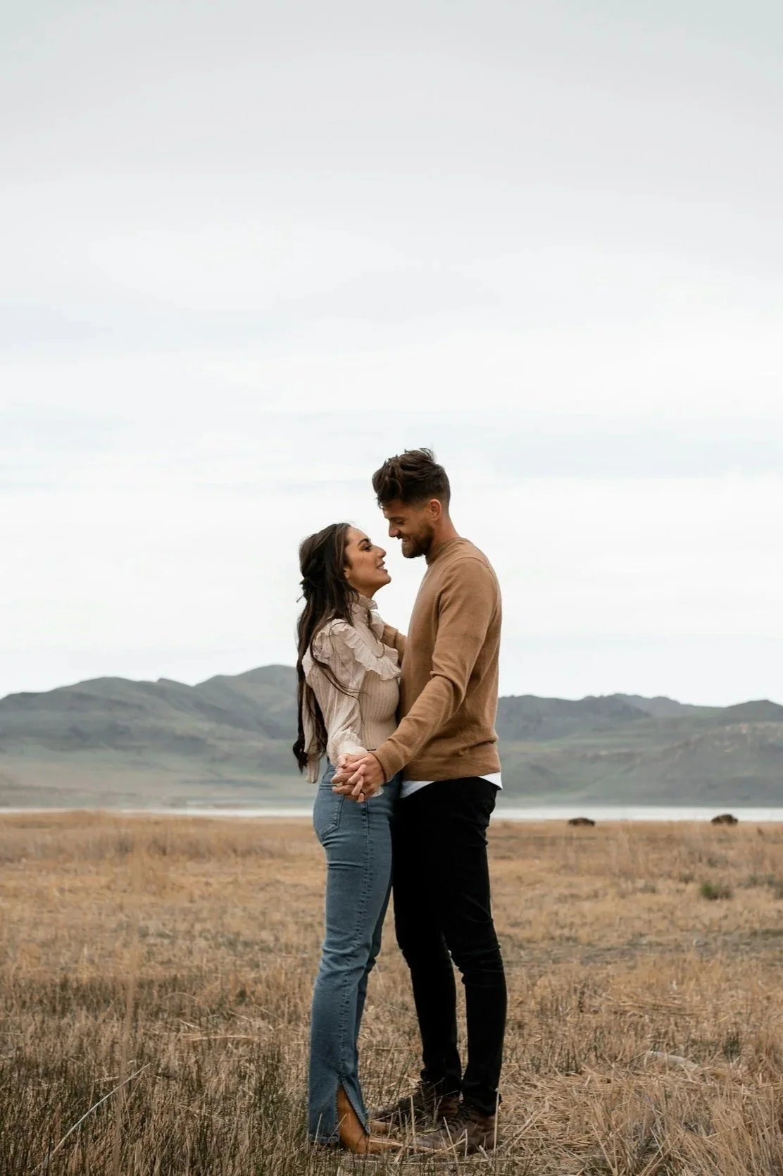 A couple stands in a field holding hands and smiling at each other with mountains in the background.