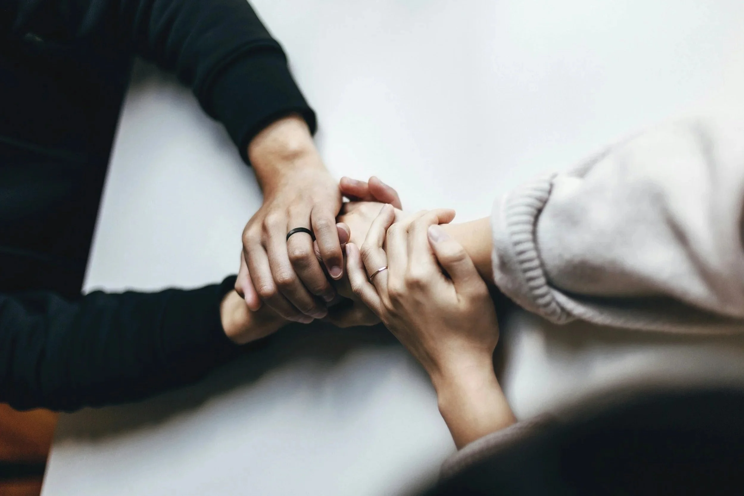 Two people holding hands, one in a black long-sleeve shirt and the other in a light-colored long-sleeve shirt, with their hands interlocked on a white surface.