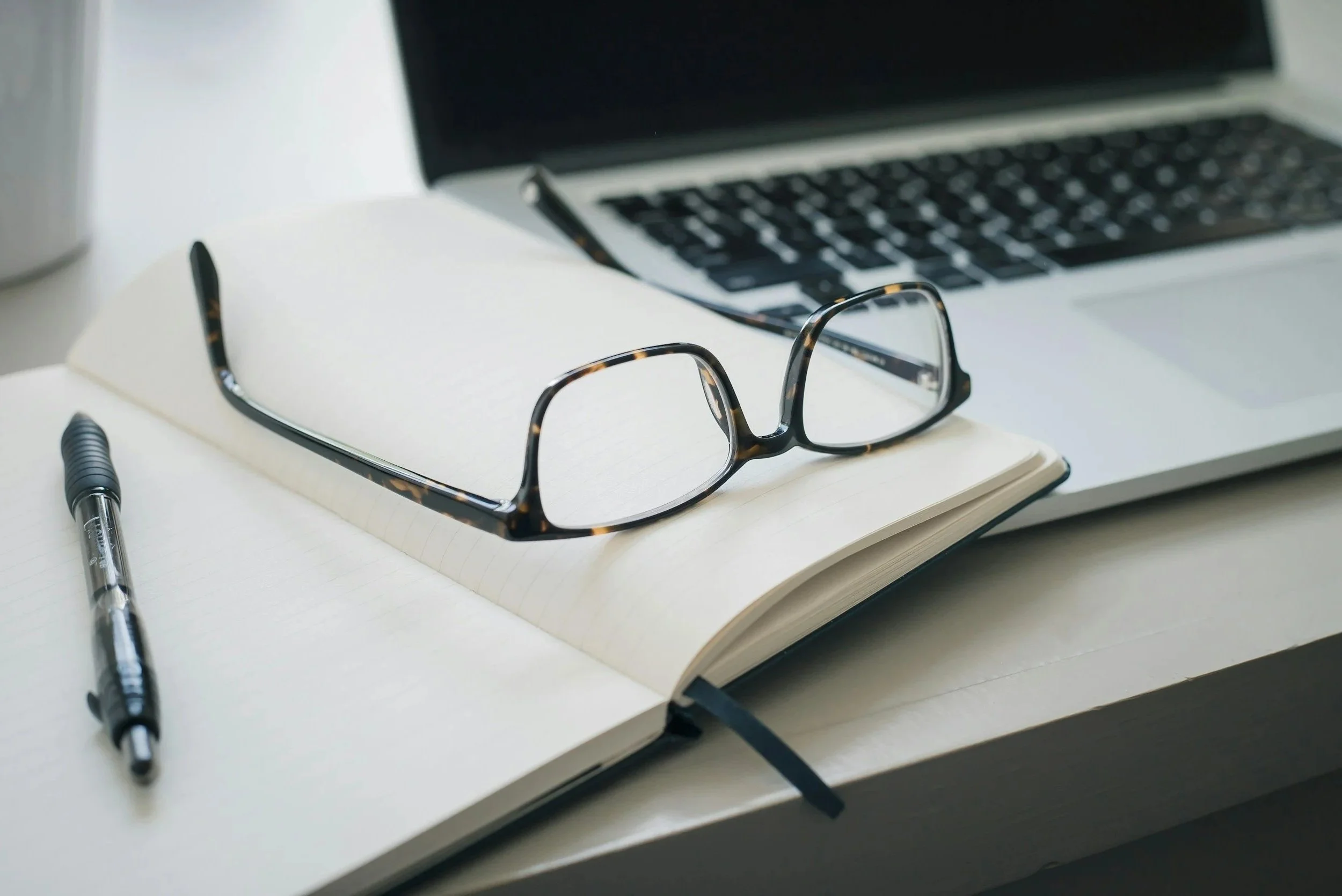 Eyeglasses resting on a closed notebook on a desk with a pen, a laptop, and a cup nearby.