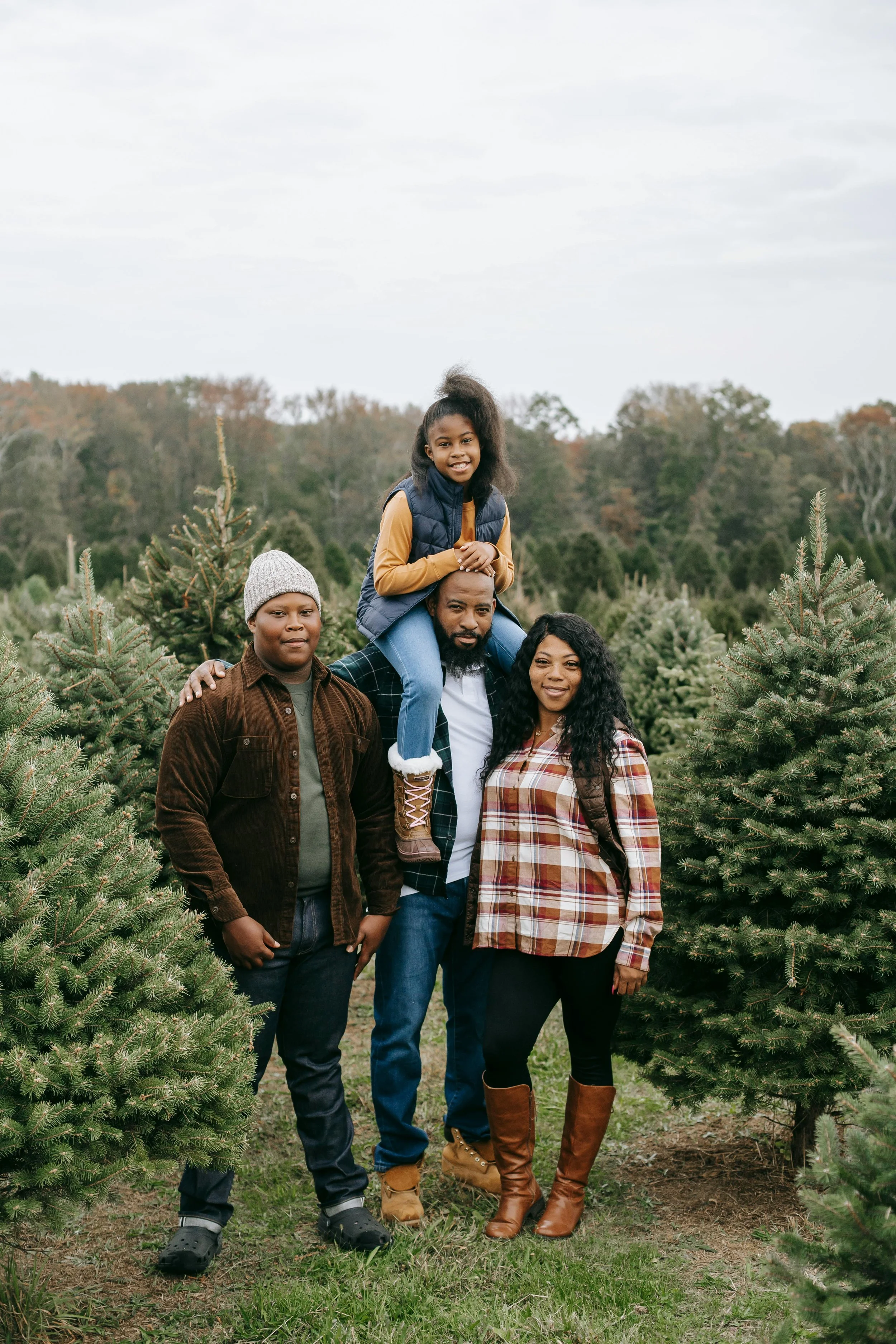 Family of four standing among Christmas trees outdoors on a cloudy day. The child is sitting on the father's shoulders, and everyone is smiling.