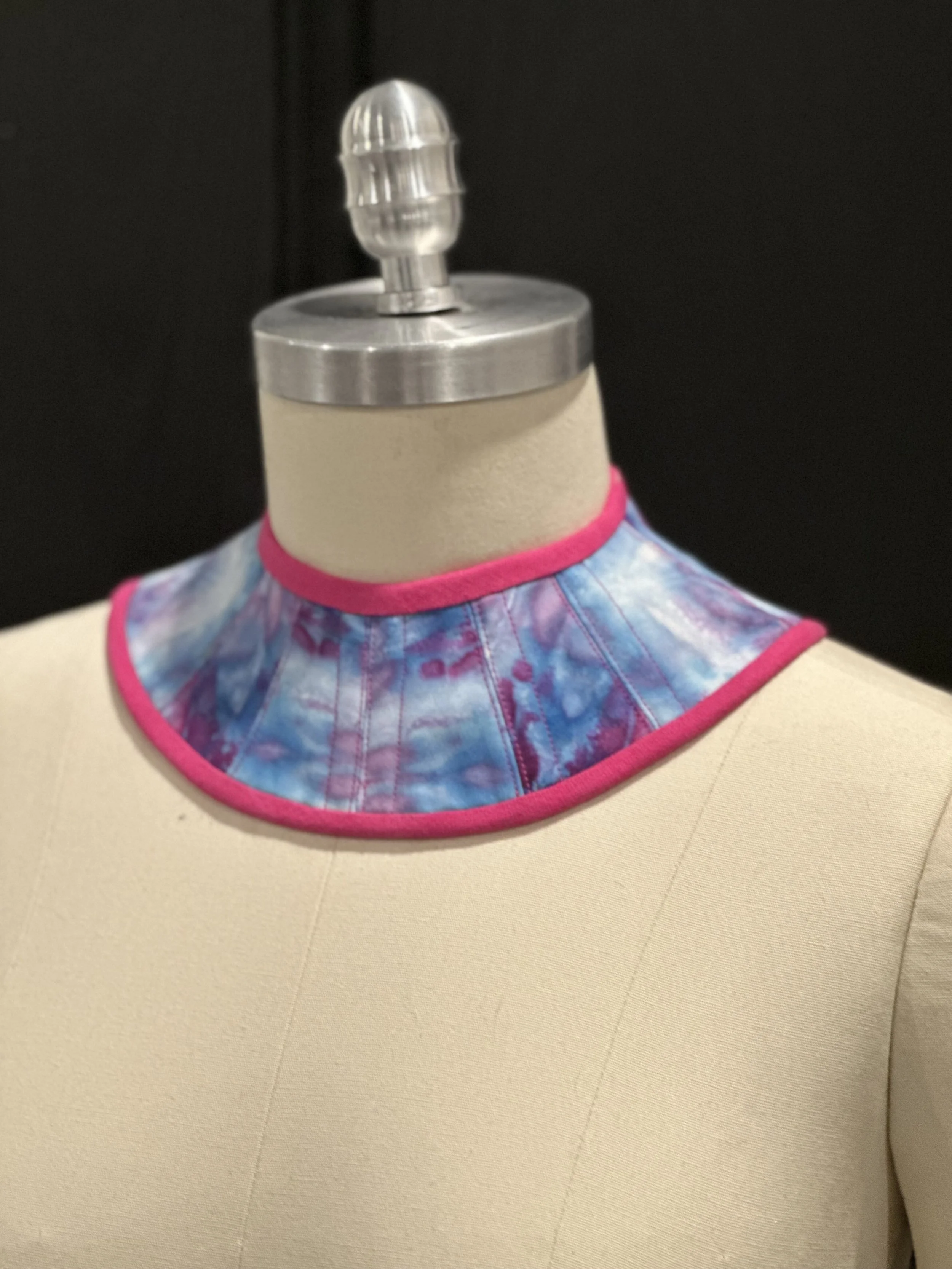 A mannequin neck displays a colorful tie-dye bib with pink piping on a beige mannequin torso, set against a black background.