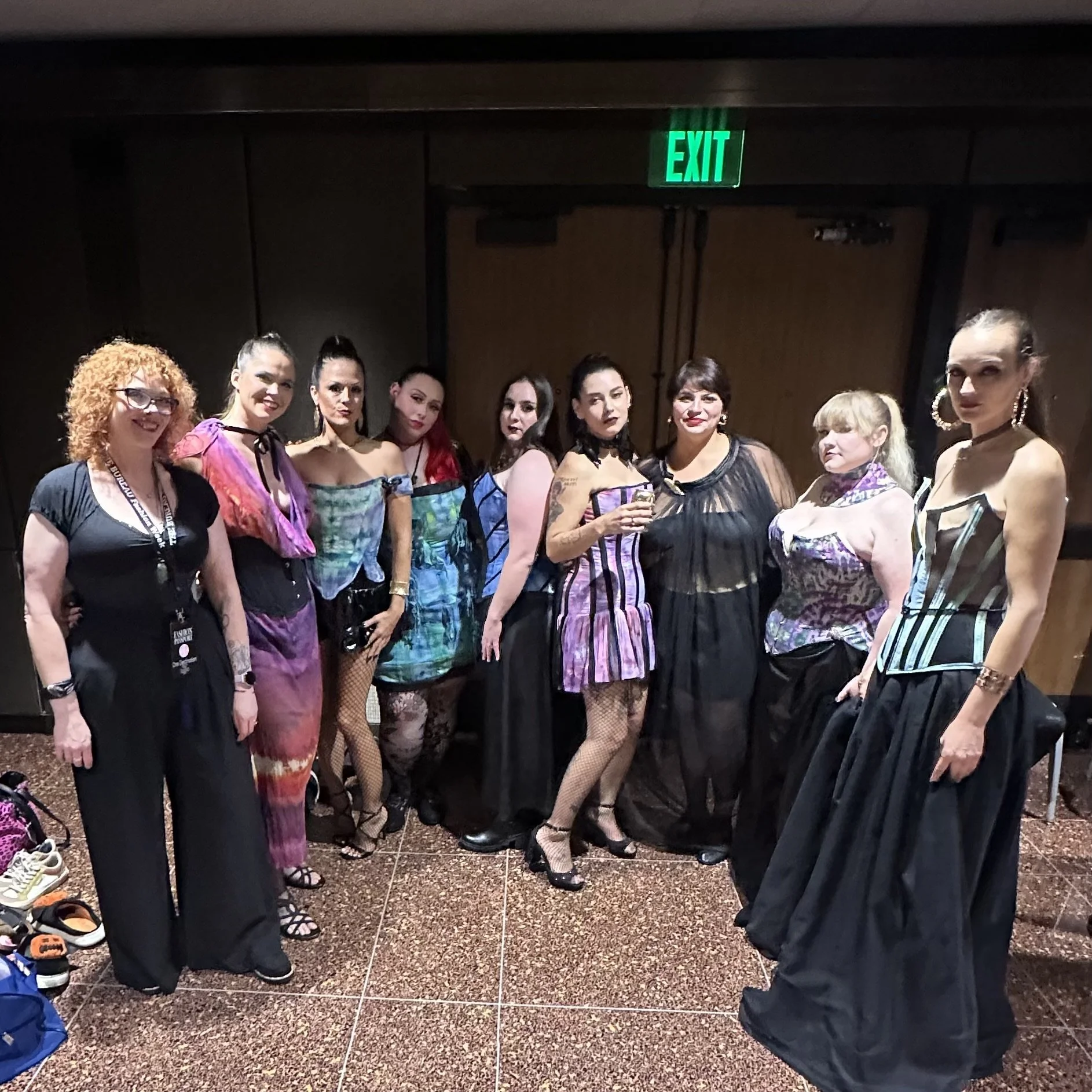 A group of models dressed in colorful, elegant corset outfits with some wearing fishnet stockings and high heels, standing together in the greenroom of a fashion show.