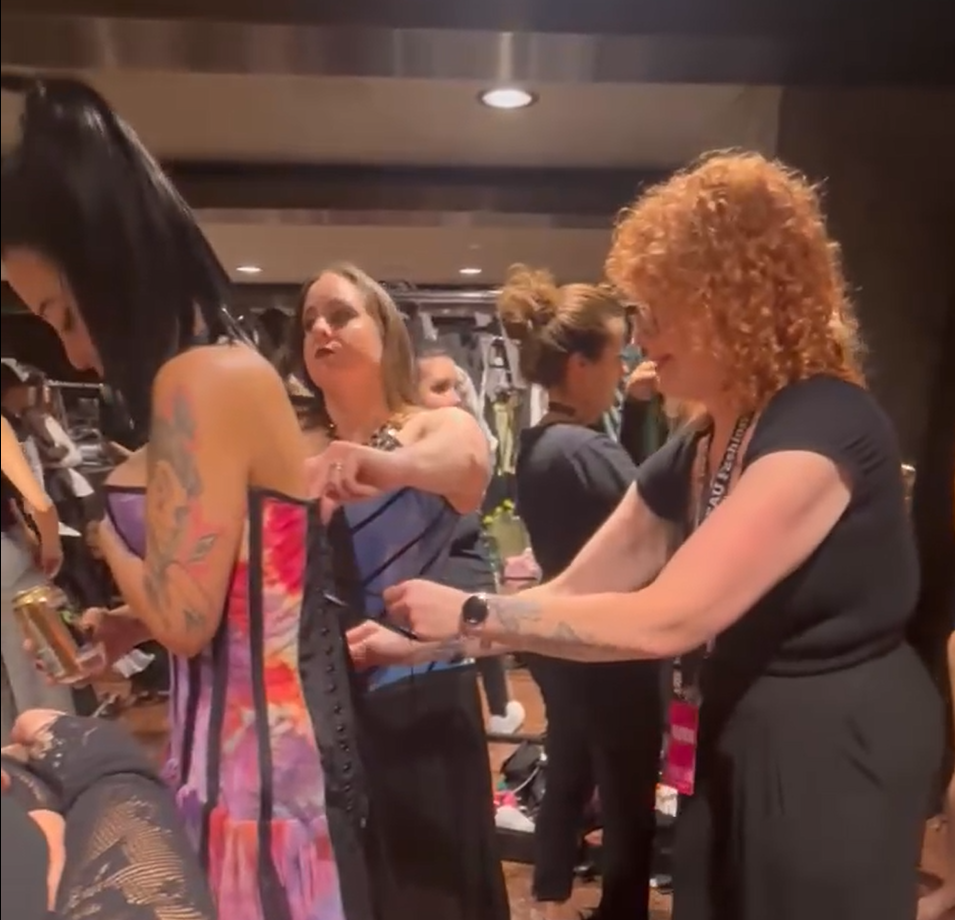 A woman with tattoos on her arm in a colorful dress standing in a crowded indoor setting, while another woman with curly red hair and a black dress is assisting her with something on her phone.