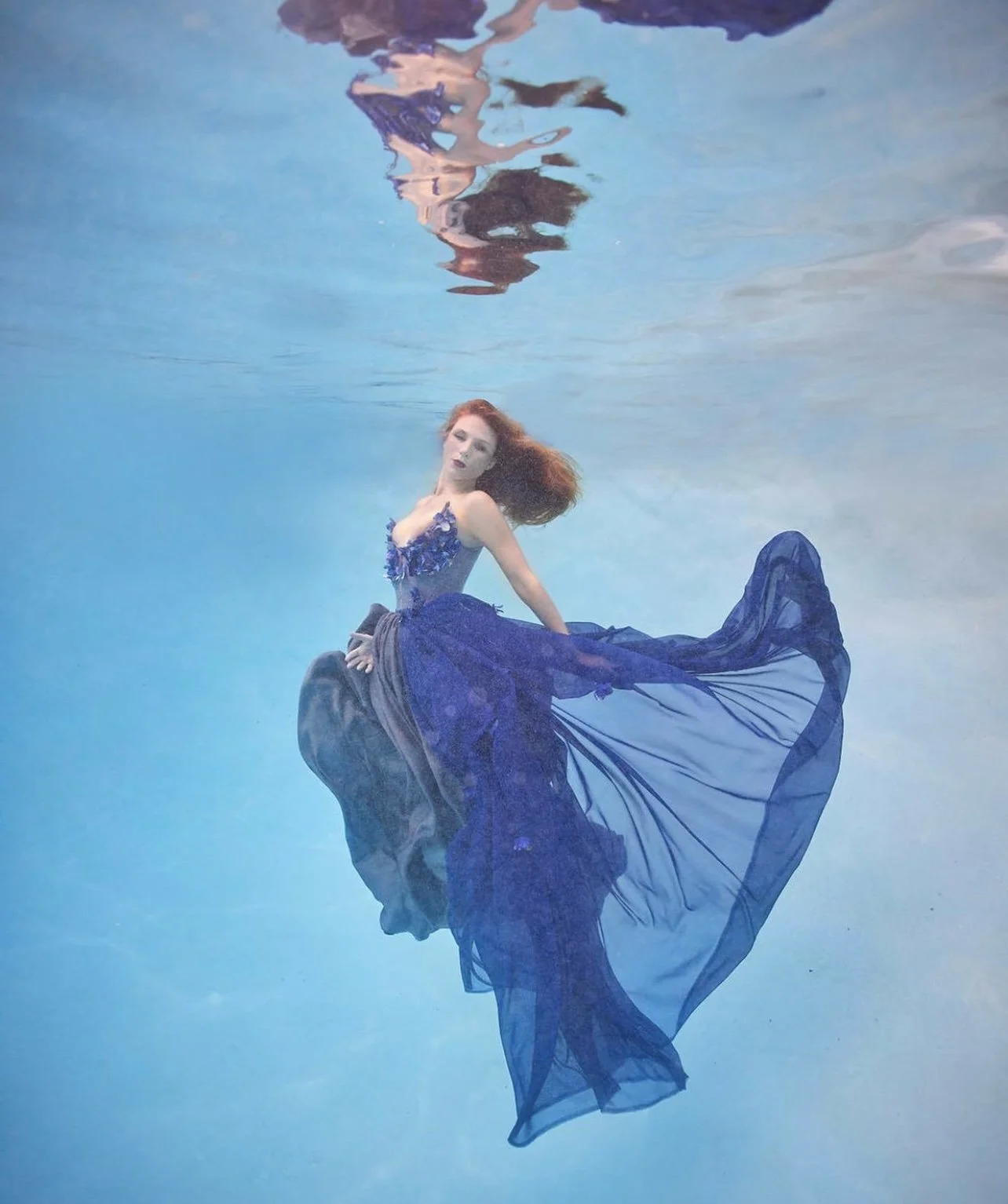 A woman with red hair in an elegant dark blue dress is underwater in a swimming pool, with her dress flowing around her.