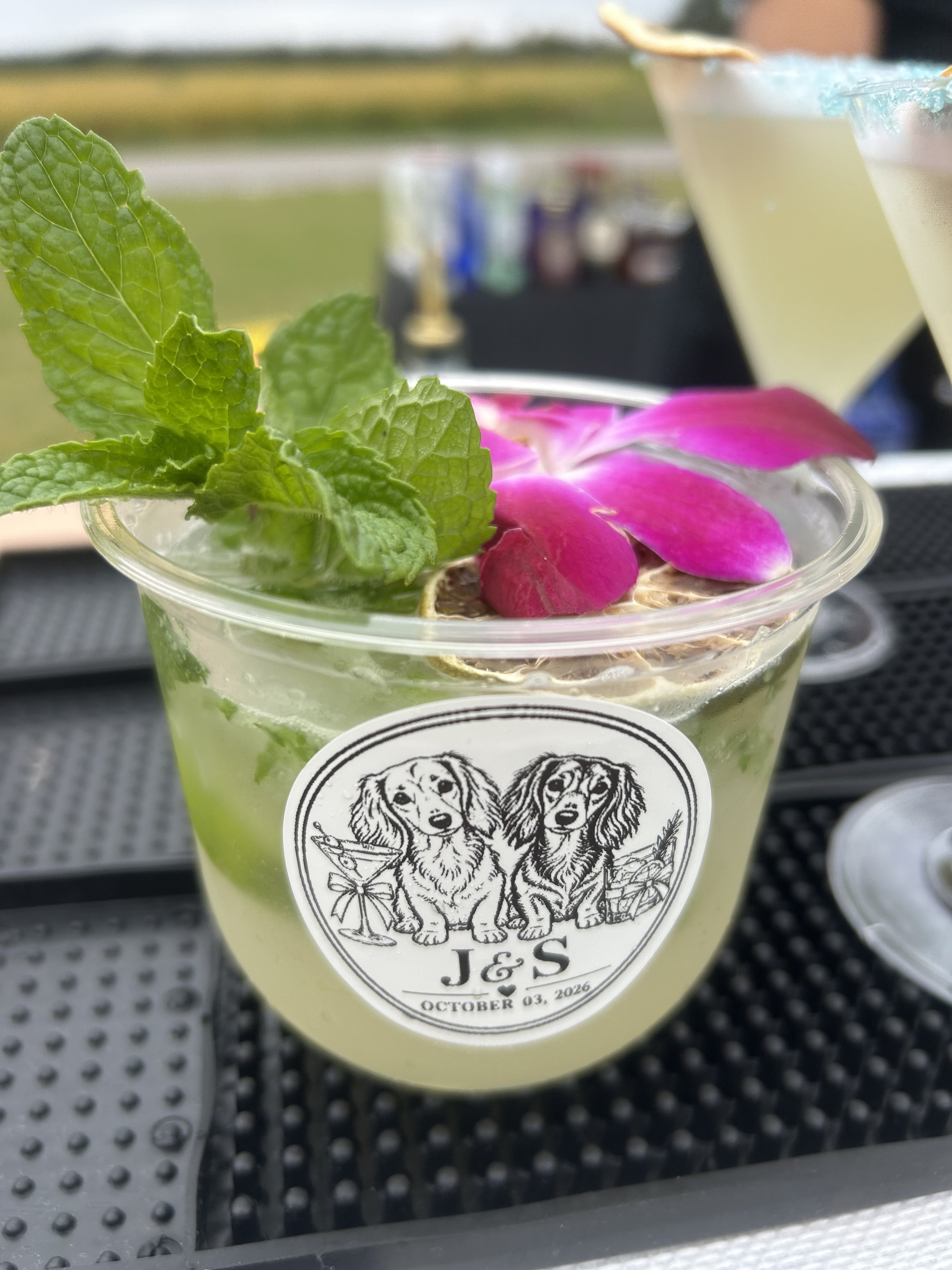 A clear plastic cup with a logo of three dogs and the text "J & S" and the date October 03, 2026. The drink inside appears to be a light-colored beverage, topped with some fresh mint leaves and pink flower petals. In the background, there are two coc