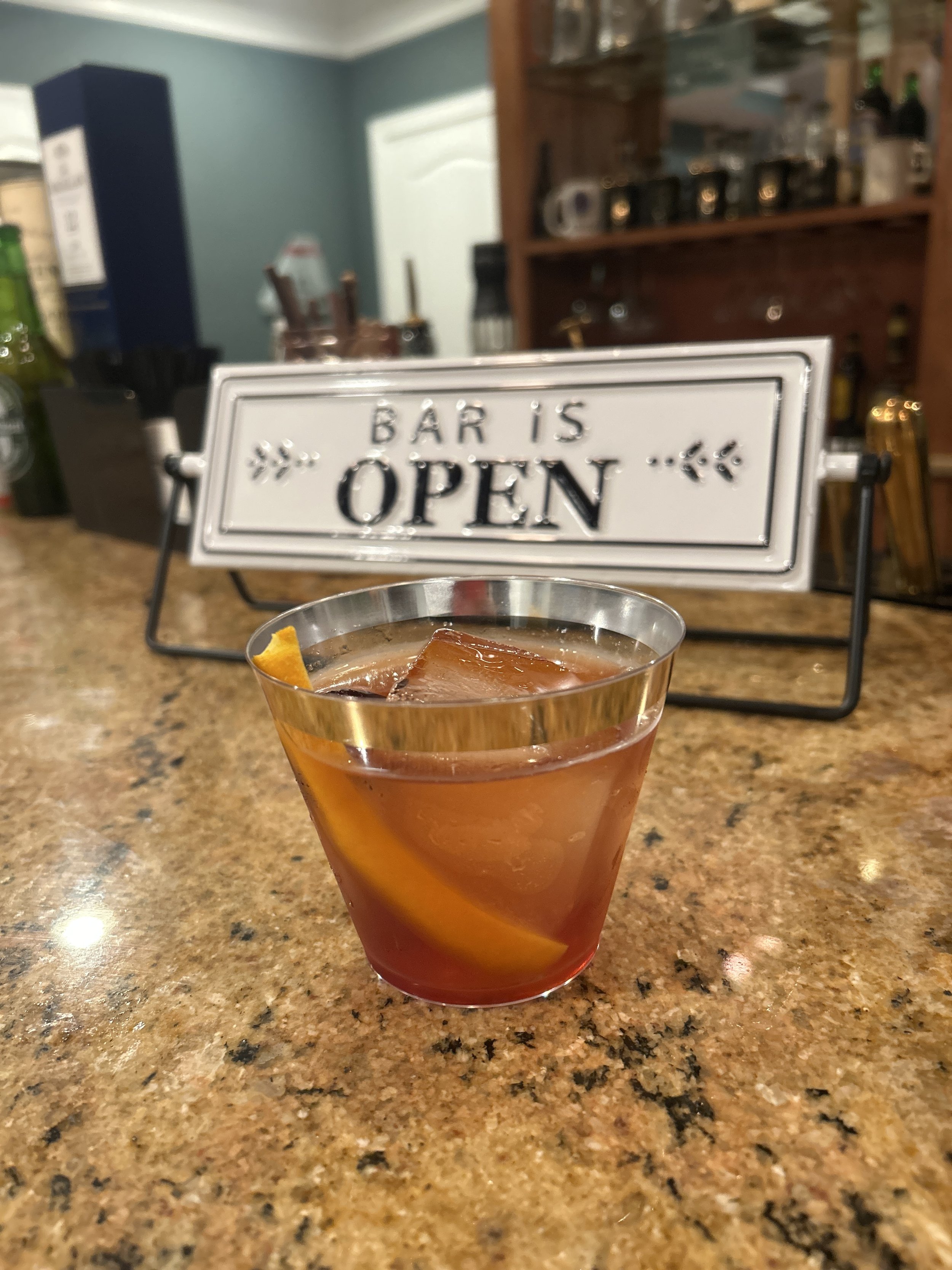 Close-up of a cocktail with an orange peel on a bar counter, in front of an 'Open' sign.