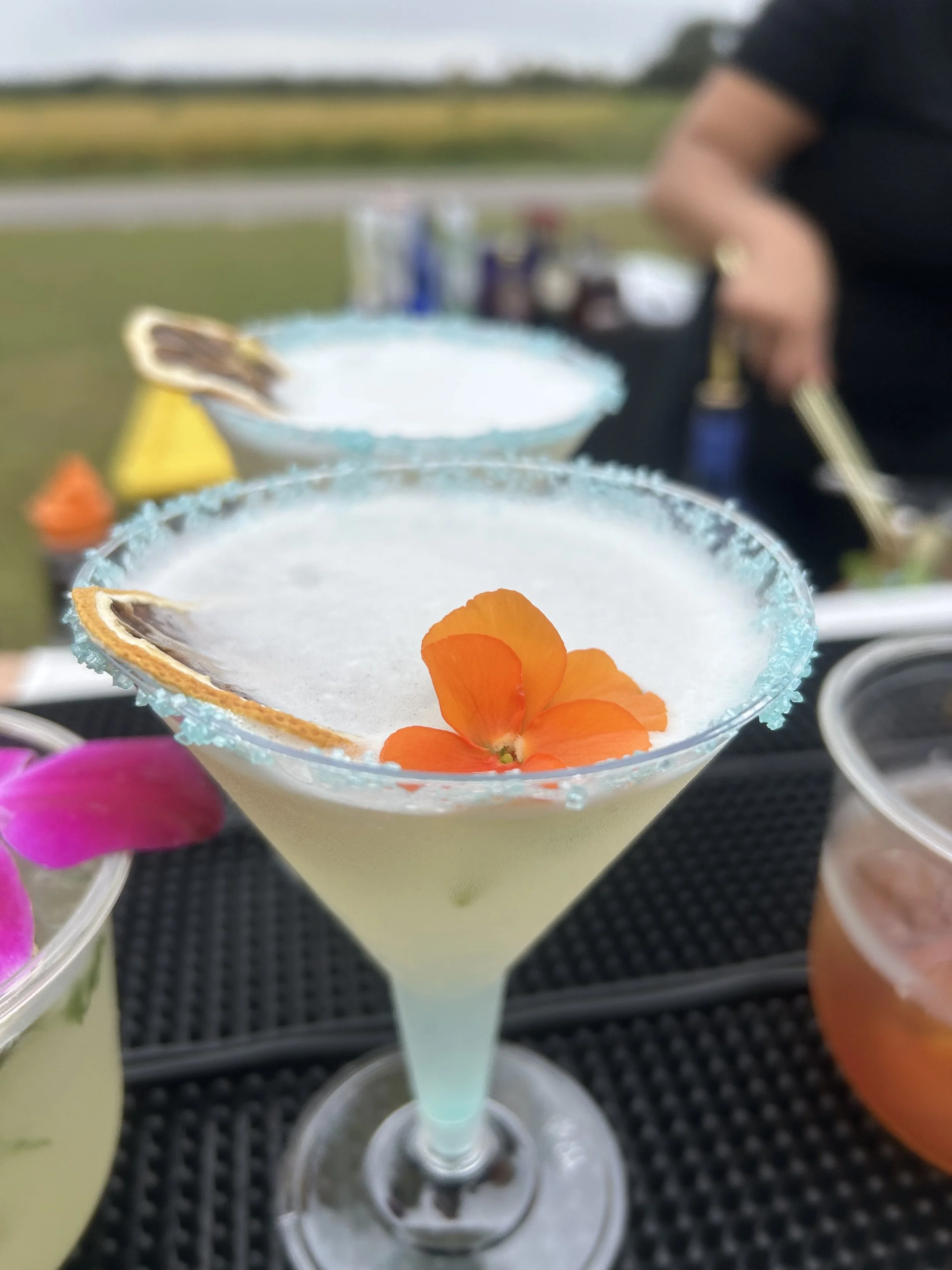 Close-up of a margarita cocktail in a classic glass with a salted rim, garnished with an orange flower, on a black textured surface, with blurred background of a person and other drinks.